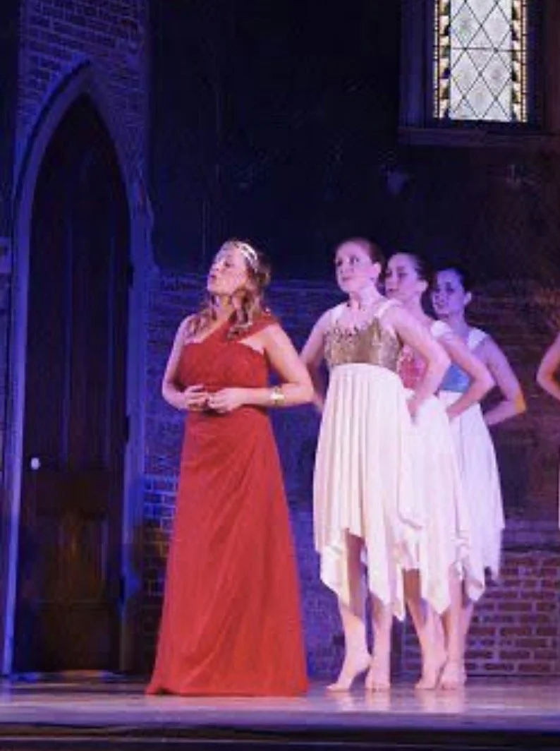 Four women performing on stage in a theatrical production, with ornate brick walls and a stained glass window in the background. One woman in a red gown is standing in front, and three women in white dresses are behind her.