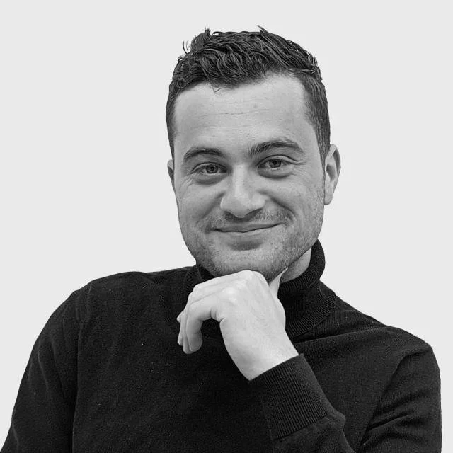 Portrait of a young man with short dark hair, smiling slightly, wearing a black turtleneck sweater, against a plain light gray background.