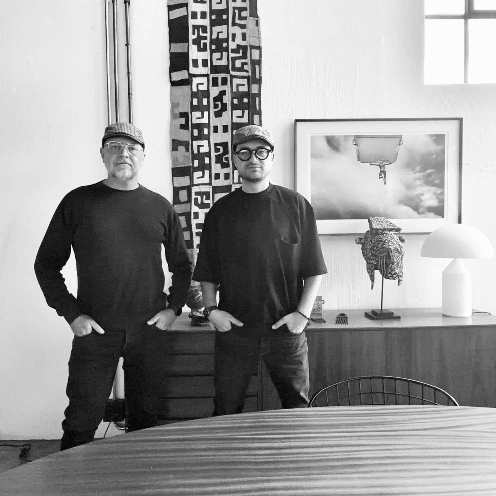 Two men stand side by side in an art-filled room. They both wear glasses, hats, and dark clothing. Behind them are decorative textiles, a framed picture of a person hanging from a basketball hoop, and various art pieces on a table and wall.