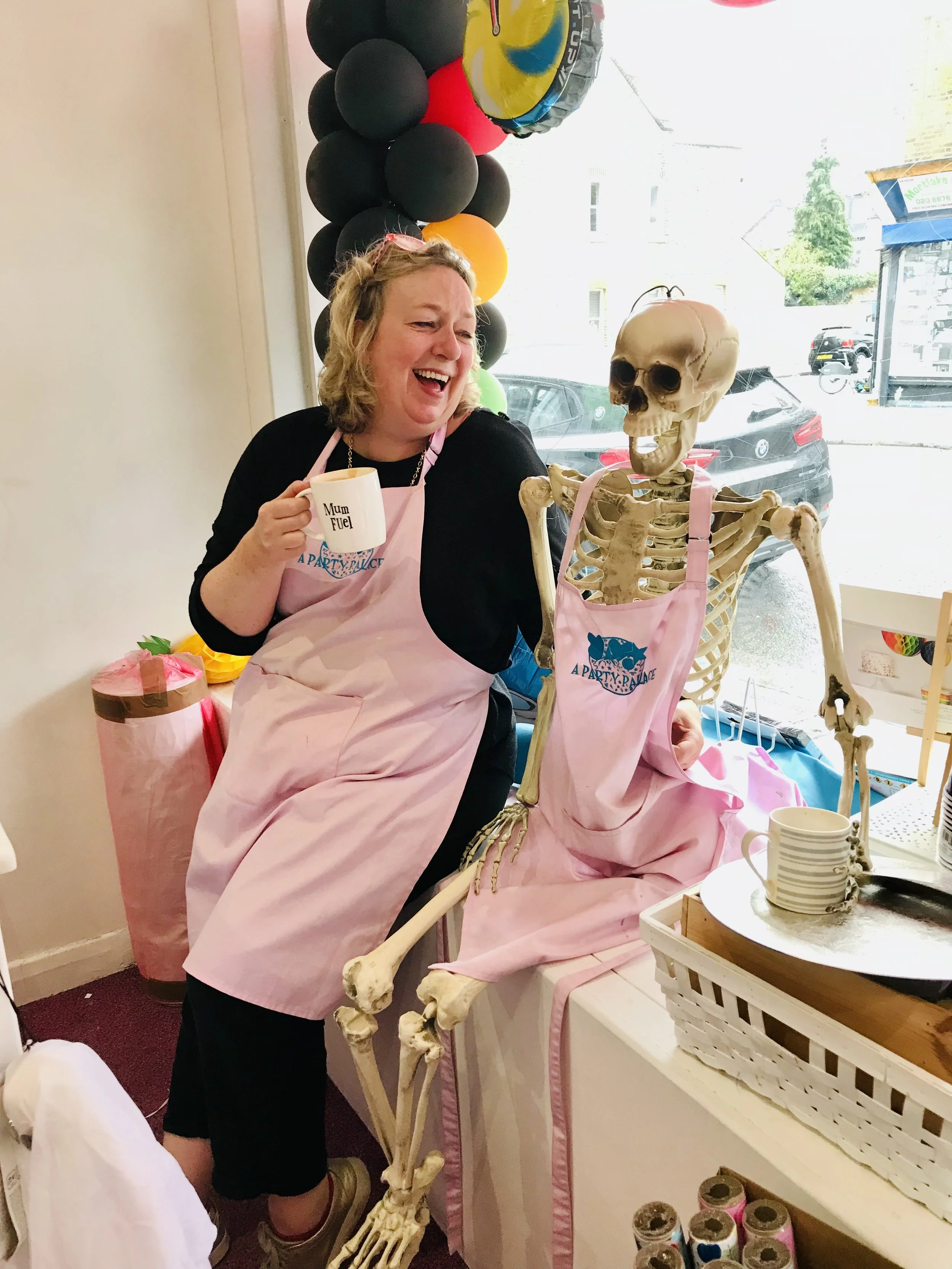 A woman is smiling and holding a mug that says 'Mum Fuel,' standing next to a skeleton. She is wearing a pink apron and glasses on her head. The scene appears to be inside a shop decorated with balloons and pink wrapping paper, with a window showing a street outside.