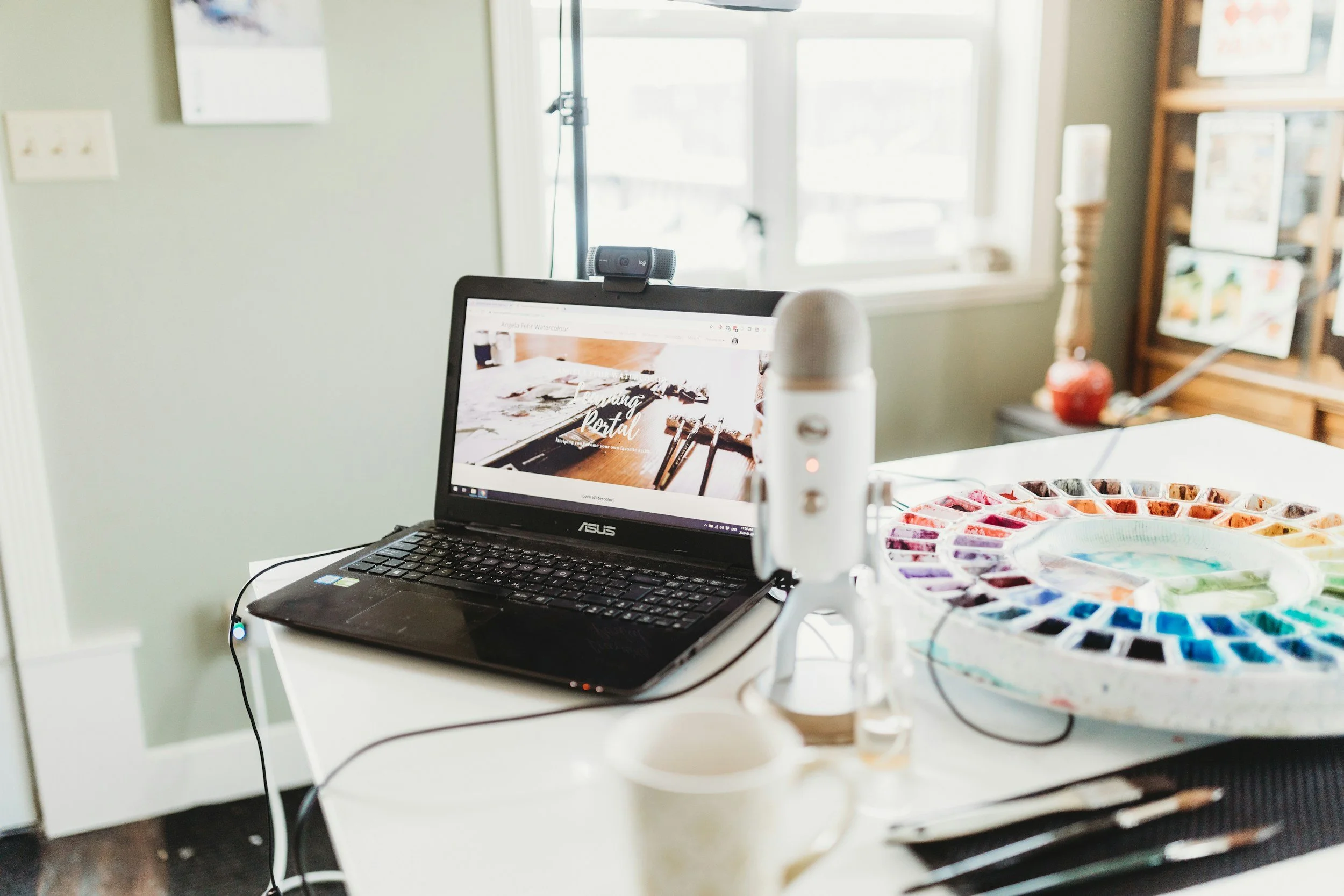 A workspace with a laptop on a white table, a watercolor paint palette, a white microphone, and a webcam, with a window and bookshelf in the background.