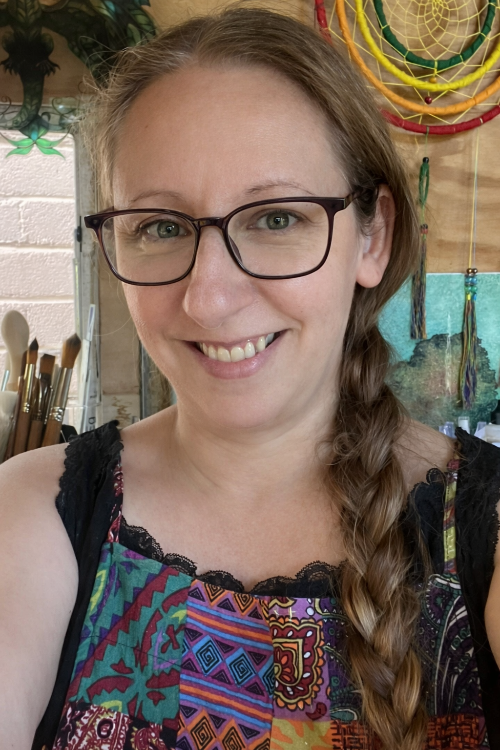 Portrait of Artist Melinda STacey of Spectrum Organising, with glasses and long braided hair, wearing a colorful patterned top, in a room with art supplies and artwork in the background.
