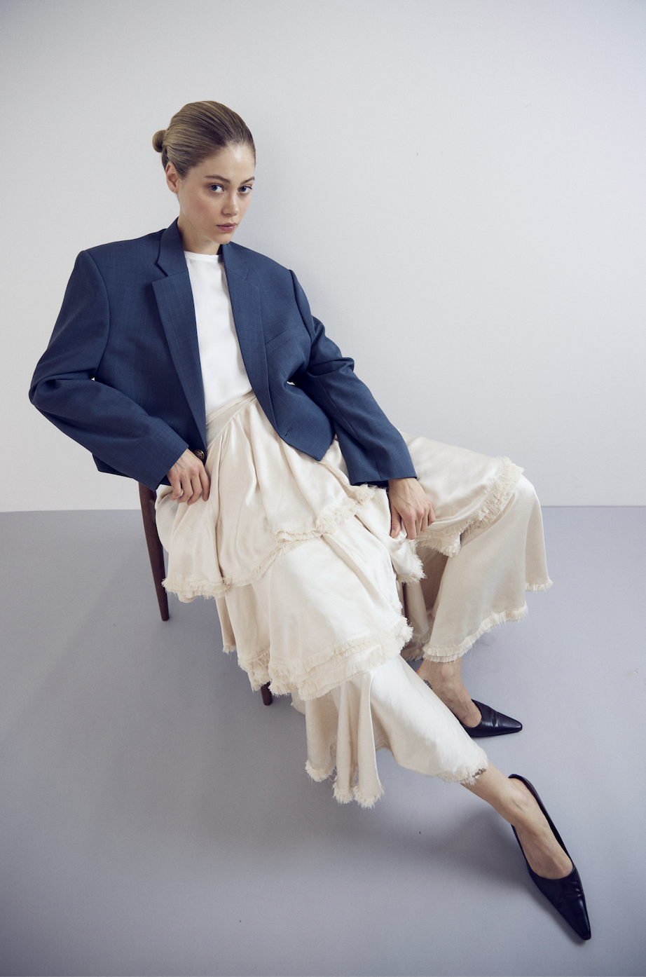 A woman sitting on a chair against a plain white wall, wearing a blue blazer, a white top, and a long, cream-colored skirt with frayed edges, paired with black pointed-toe heels.