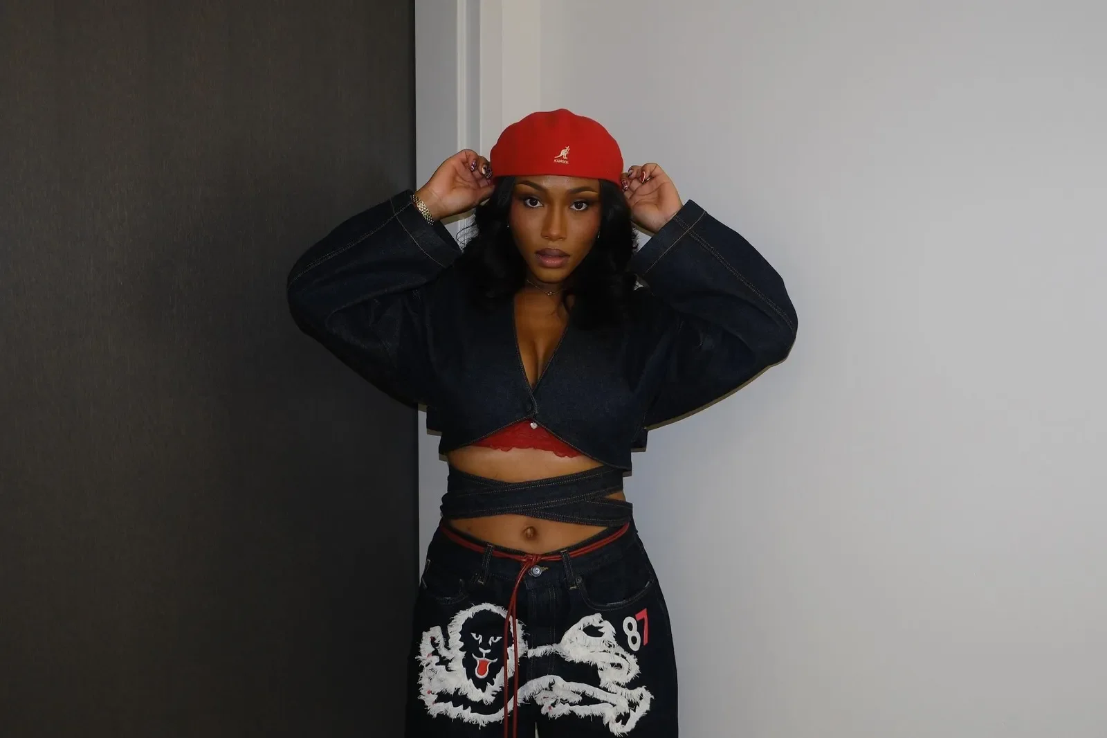 A woman with dark hair standing against a background featuring a dark wall and a light wall, wearing a red cap, a black cropped jacket, a red bralette, and black pants with a white lion graphic and the number 87, adjusting her cap with both hands.