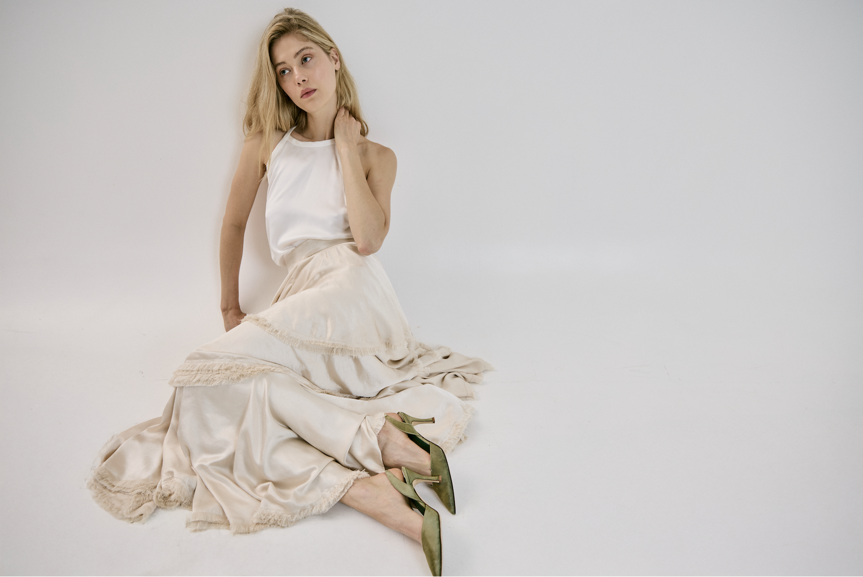 Young woman with blonde hair sitting on the floor against a plain white background, wearing a white satin top, a beige layered skirt, and green high heels.