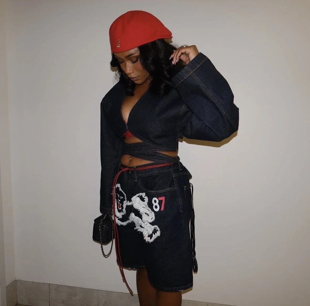 A woman with dark hair wearing a red beanie, black denim jacket, black shorts with a tiger and the number 87 embroidered on them, and a small black purse with a chain strap, standing against a plain wall.