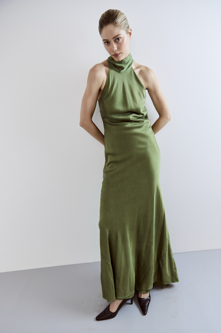 A woman with blonde hair styled in an updo, wearing a sleeveless olive green satin gown with a high neck, standing against a plain light-colored background.