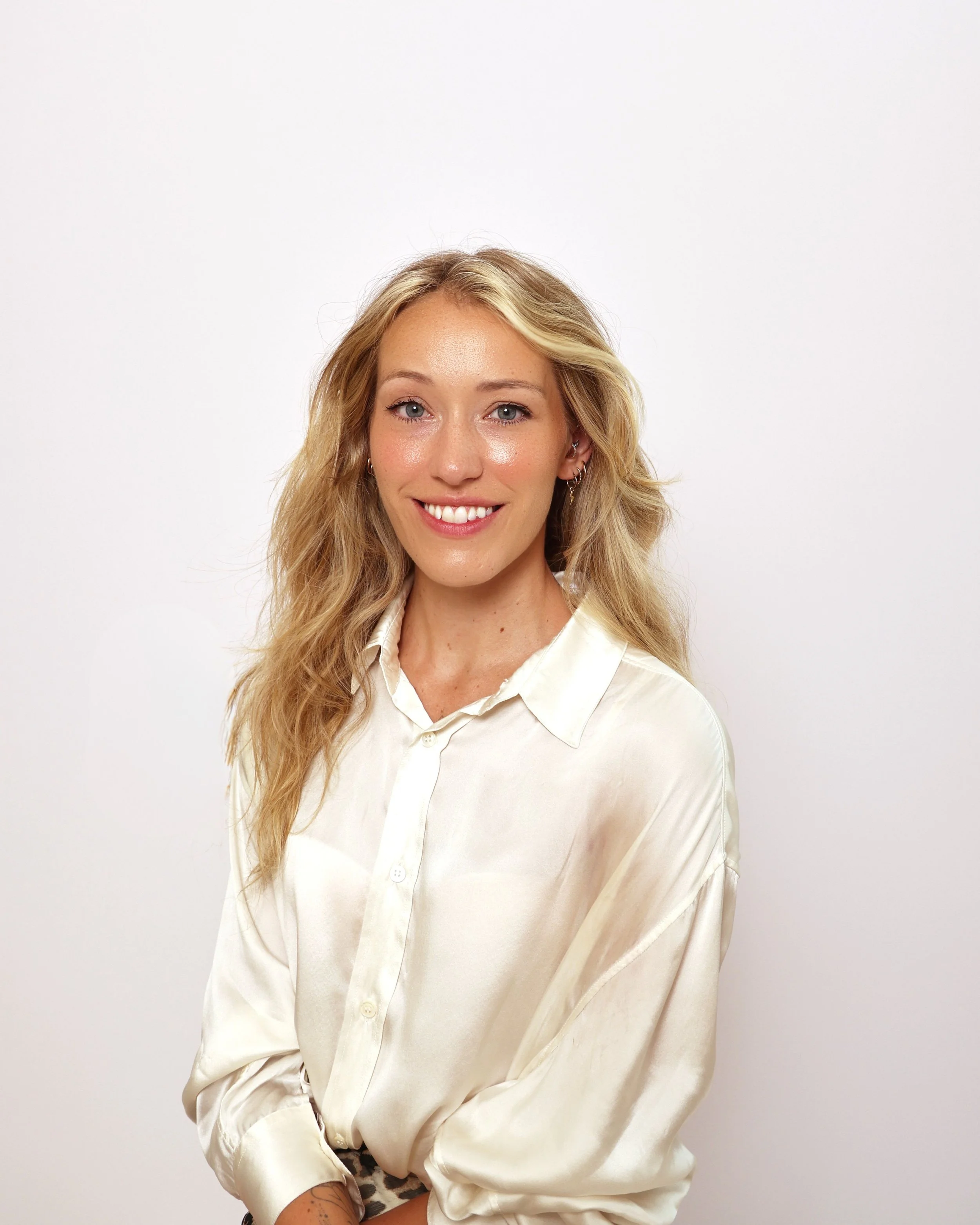 A woman with long, wavy blonde hair, blue eyes, and light makeup, wearing a cream-colored satin blouse with a pointed collar and pearl buttons, smiling in front of a plain white background.
