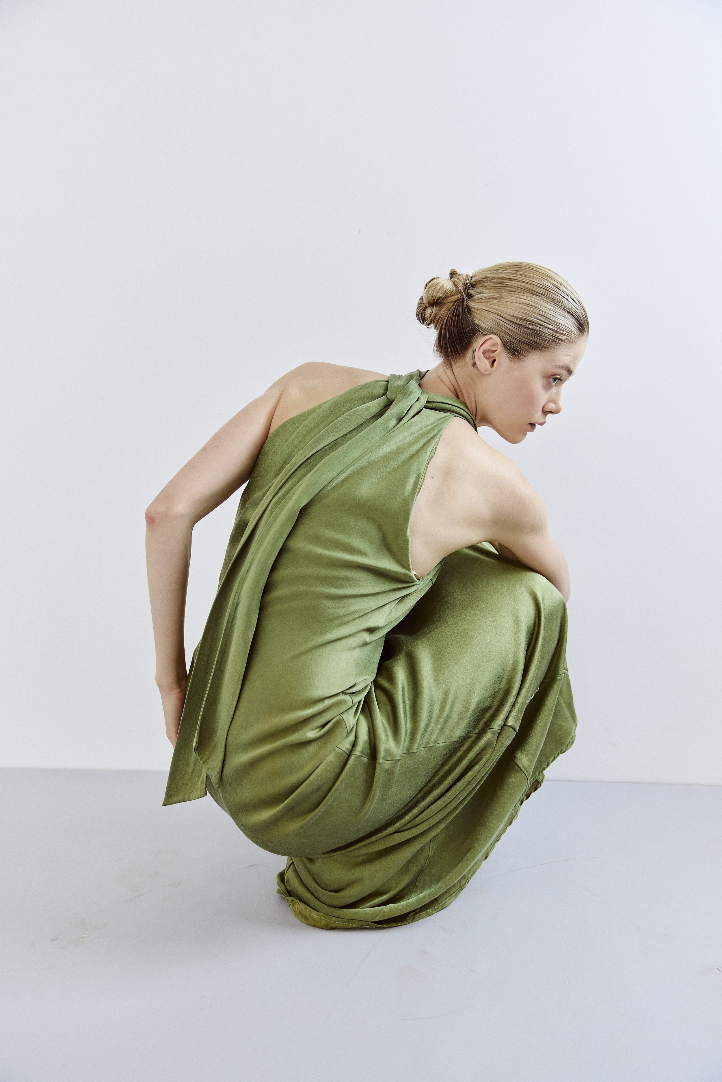 A woman dressed in a loose, flowing olive green outfit, crouching on the floor with her right knee bent and left knee near her chest, her hair styled in a slicked-back bun, against a plain white background.