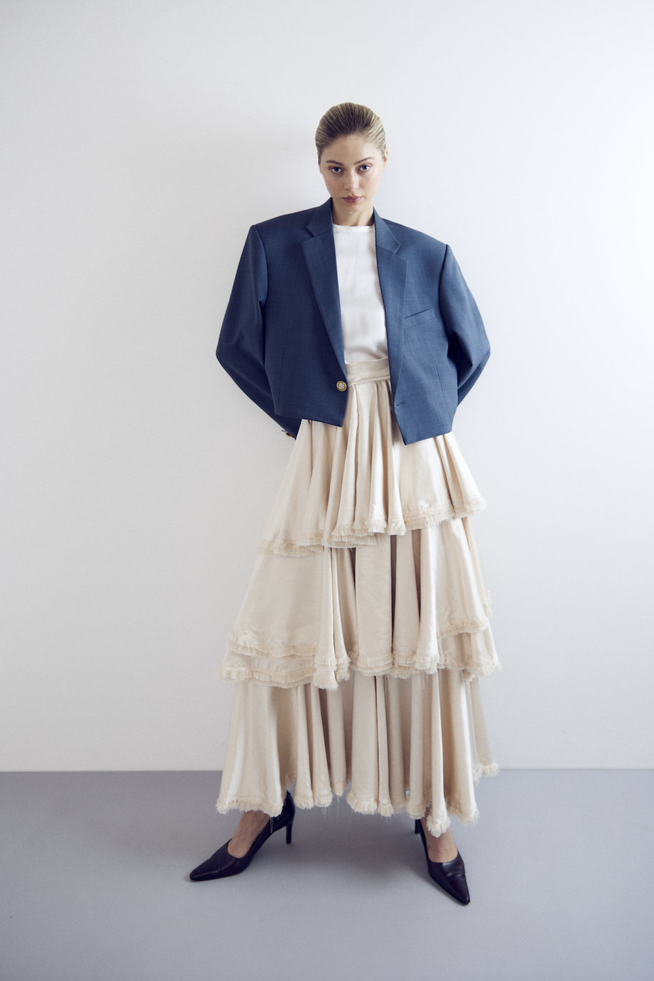 Woman standing against plain white wall, wearing a blue blazer, white shirt, beige layered skirt, and black high heels.