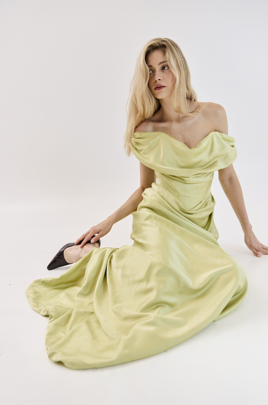 Woman with long blonde hair in a light yellow satin off-shoulder evening gown sitting on the floor, against a plain white background.