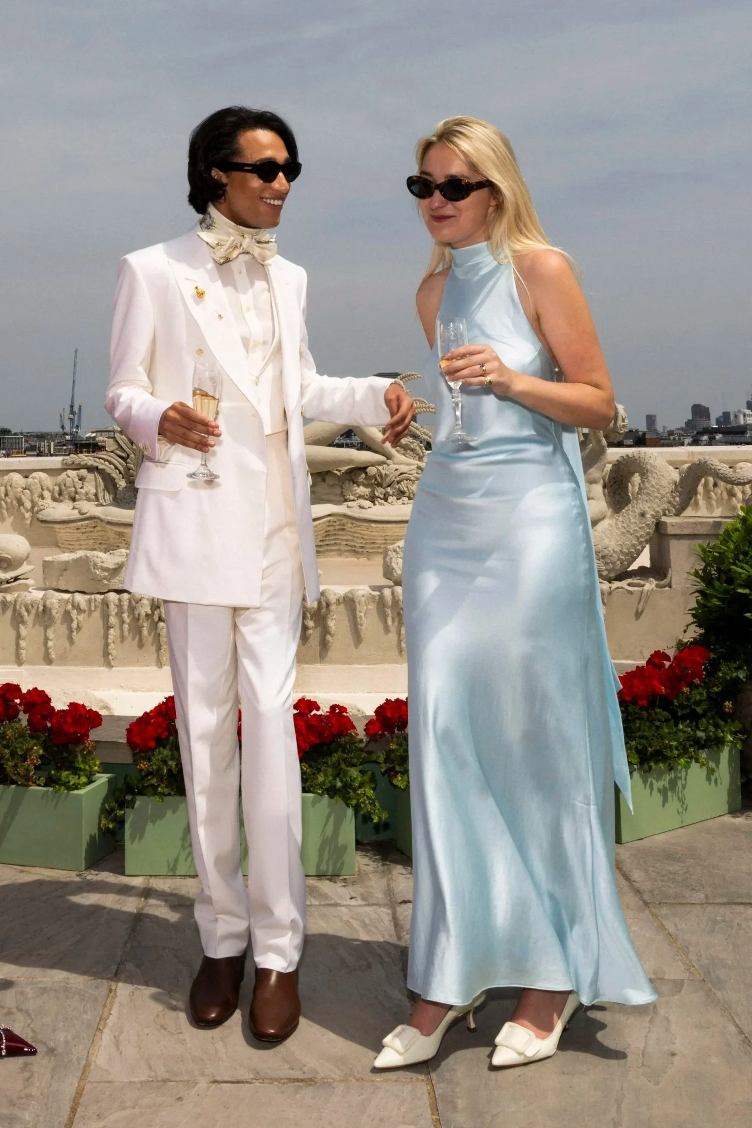A man and woman at a rooftop terrace, dressed elegantly, holding glasses of champagne, with city buildings in the background and red flowers in green planters in the foreground.