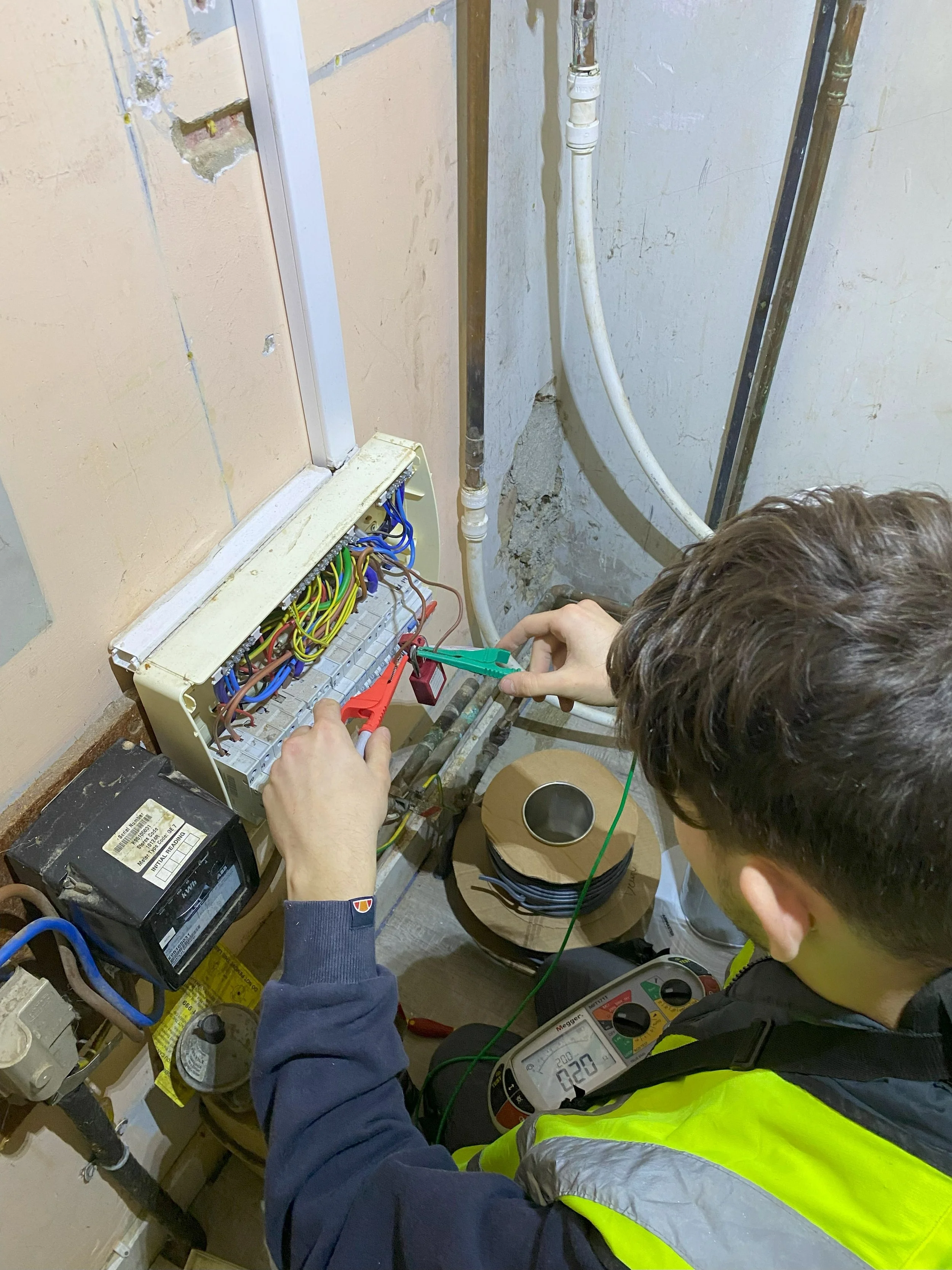 Two electricians are working on an electrical box with colorful wires, using testing equipment and tools. The scene appears to be in an unfinished room with exposed drywall and piping.