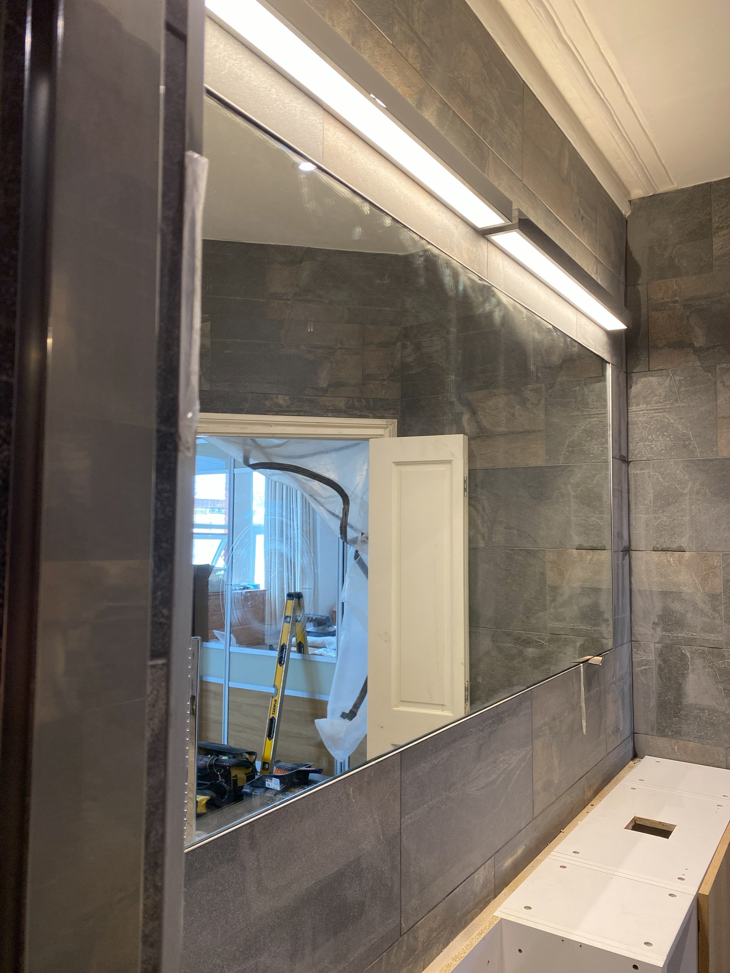 A large mirror being installed on a tiled wall in a bathroom. The mirror reflects a window and some construction tools, including a ladder and a power drill. The wall has dark gray tiles, and there is a white cabinet or vanity base in front of the mi