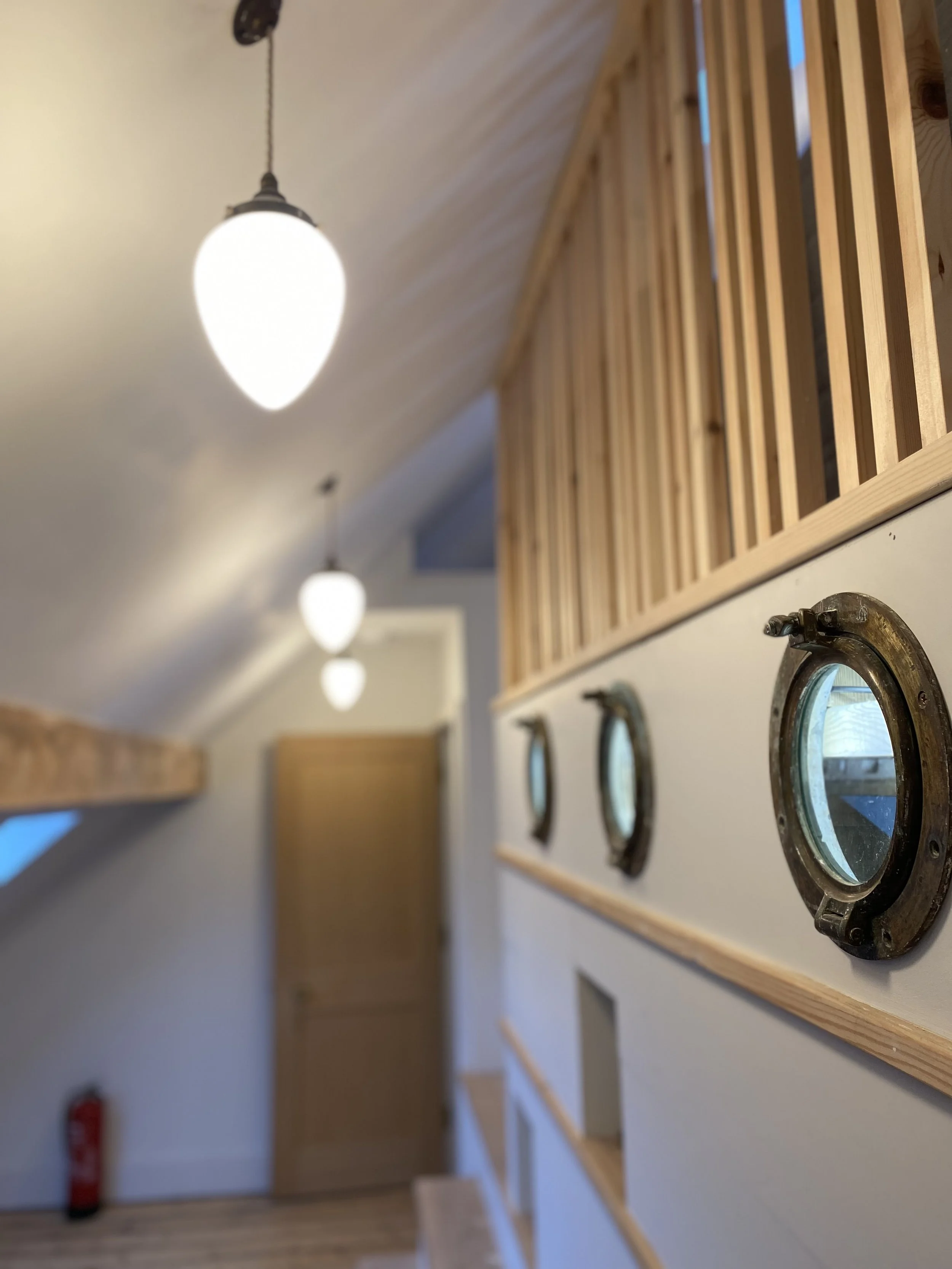 Interior of a building with a hallway wall featuring small porthole-style windows with metal frames and wooden trim, along with hanging light fixtures.