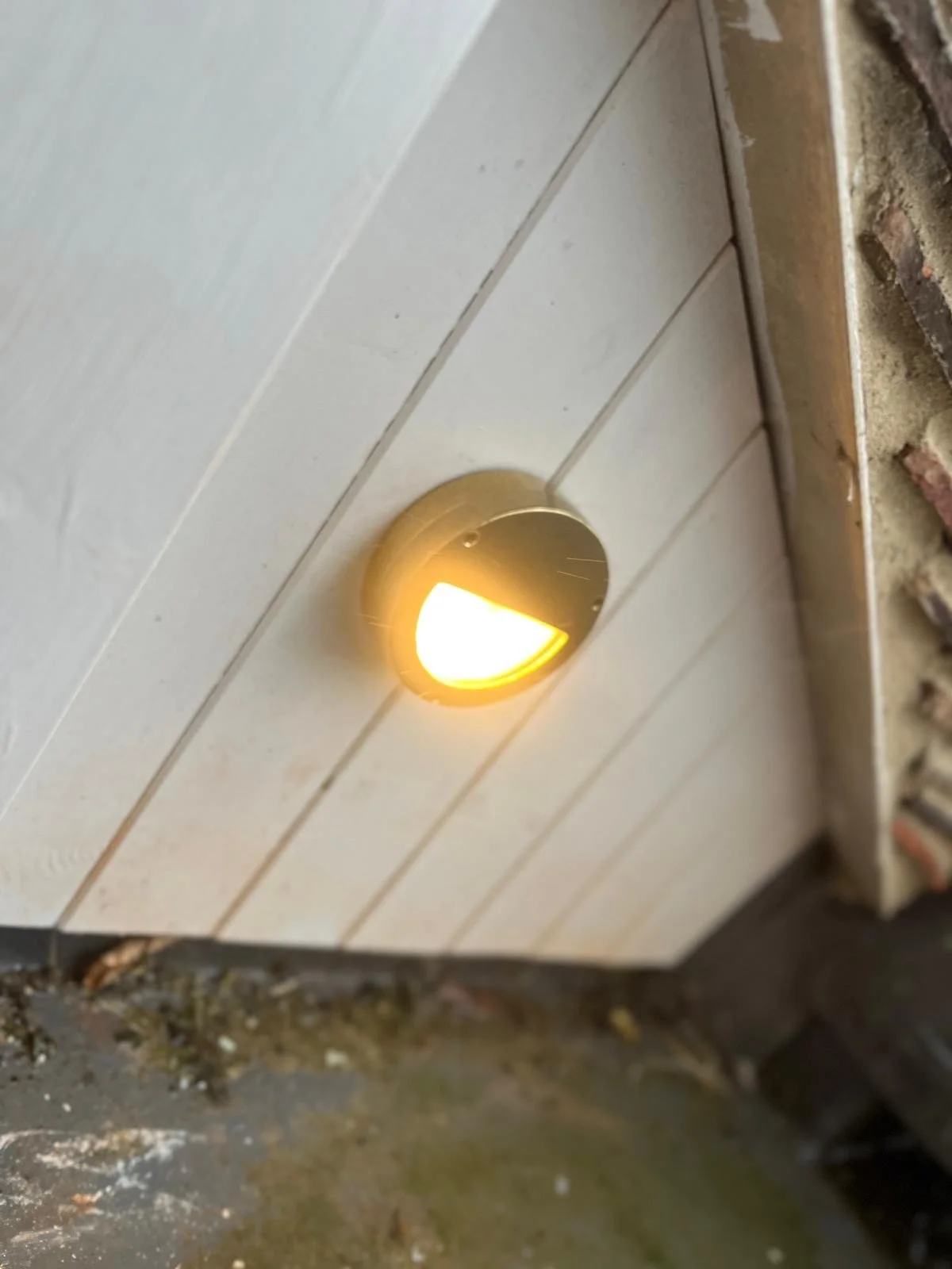 Outdoor Lighting: Close-up of a modern exterior wall light fixture emitting warm yellow light, mounted on a white wooden panel wall near a brick ledge.