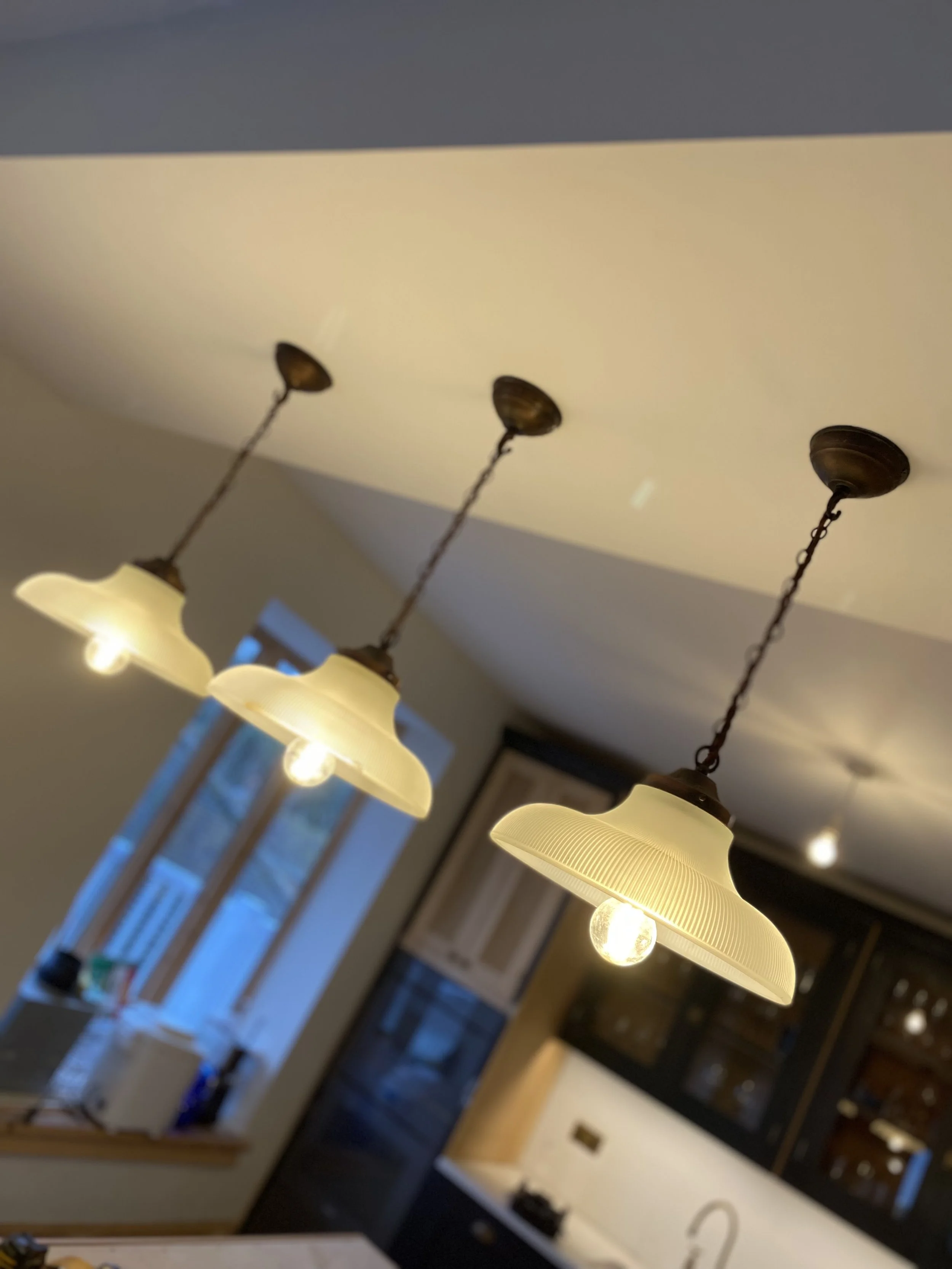 Multiple vintage-style hanging pendant lights with cream-colored shades in a room with wooden floors and a large window in the background.