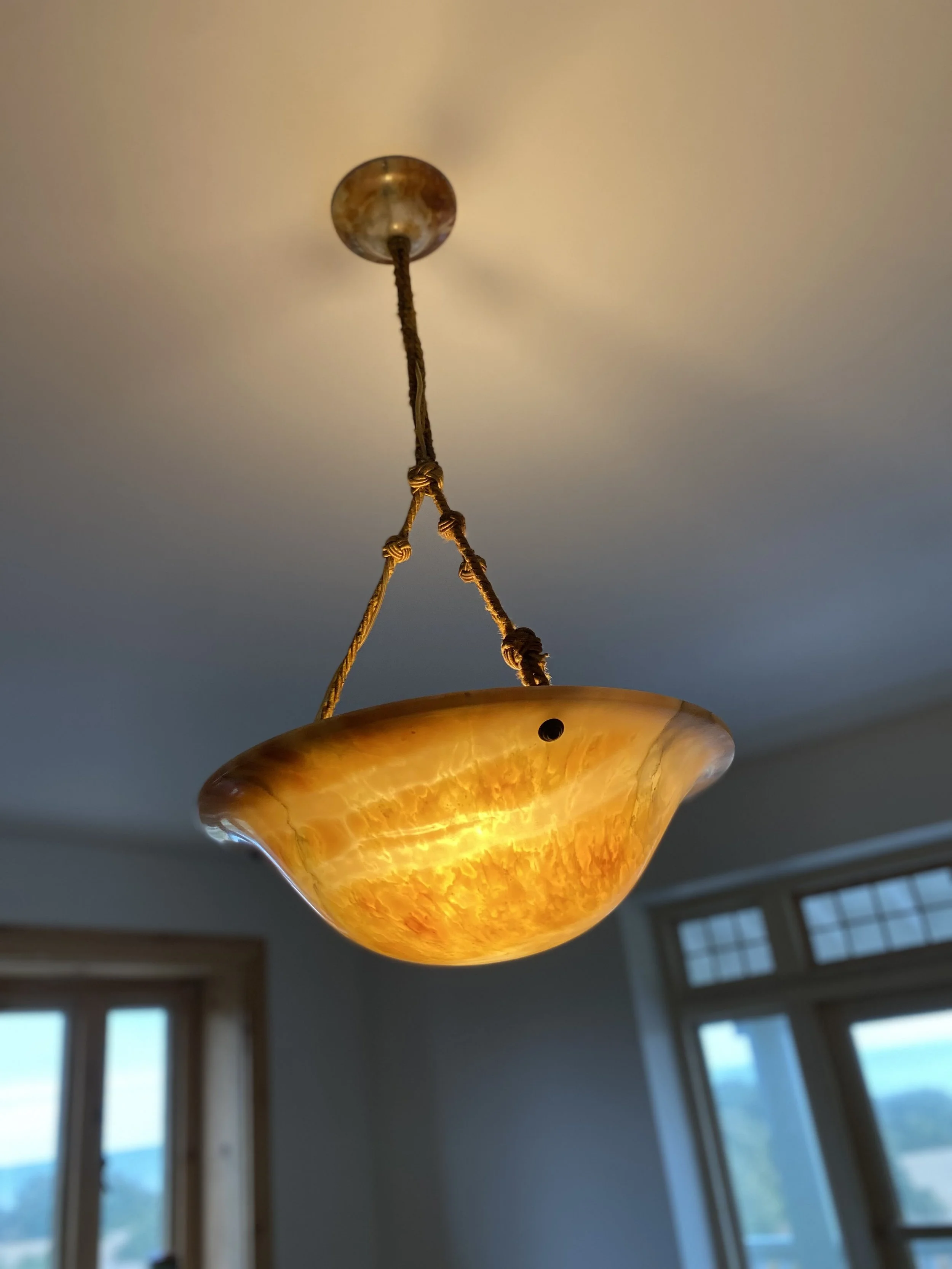 Close-up of a ceiling light fixture with a yellowish-orange glass shade, hanging from a ceiling with dark brown decorative cords, in a room with large windows and a view of mountains outside.