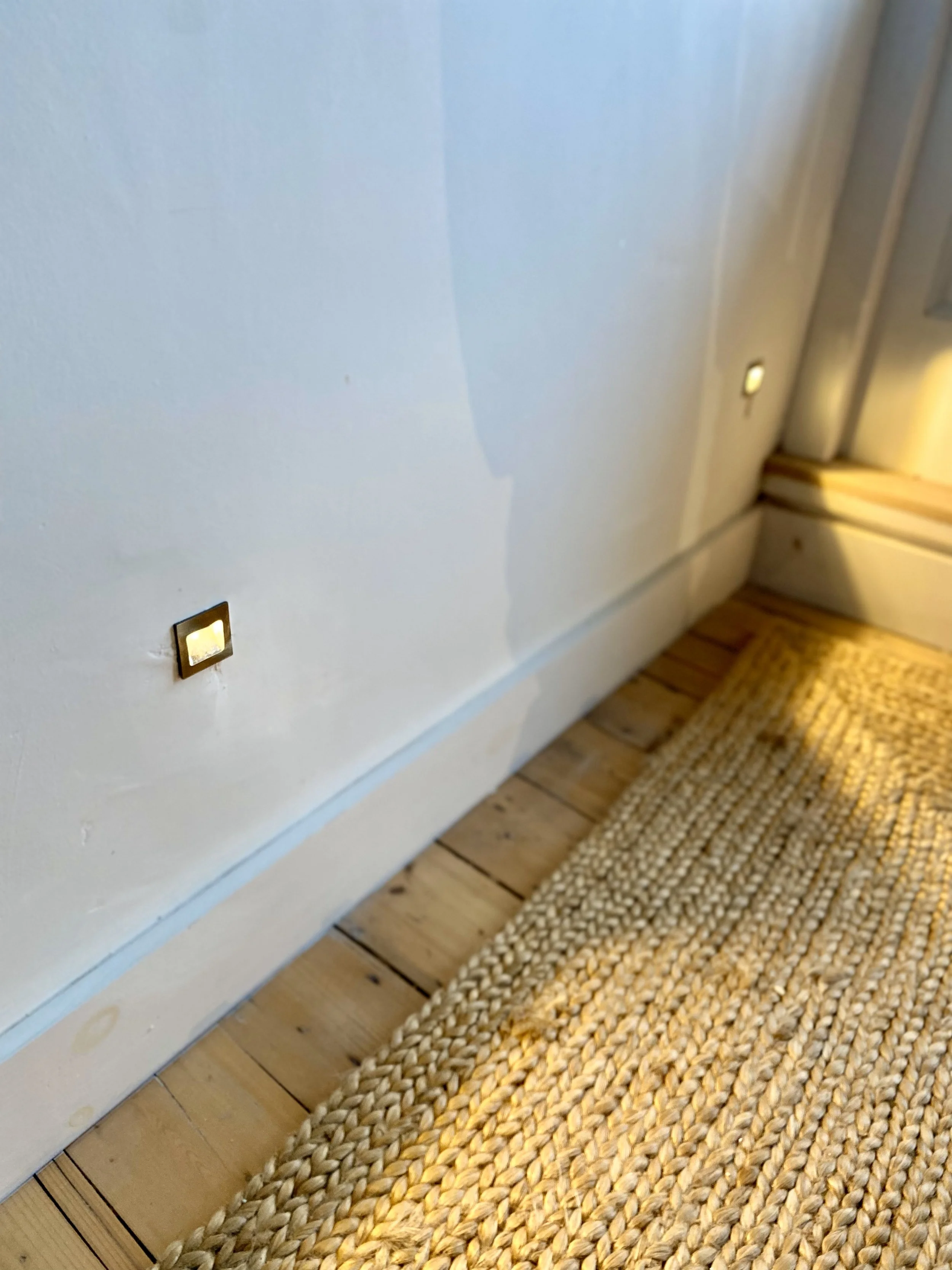 A close-up of a white wall with three electrical outlets near the floor, adjacent to a woven rug and wooden flooring.