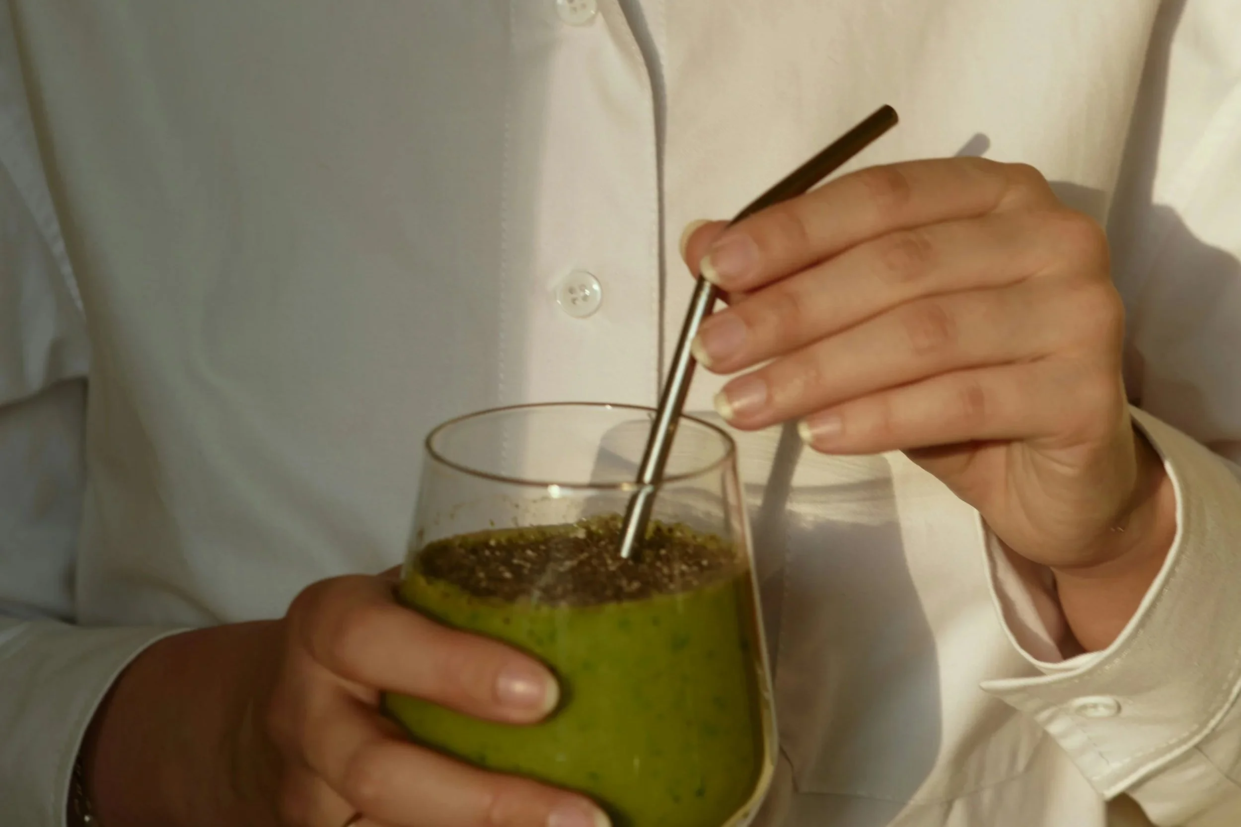 Une personne en chemise blanche tenant un verre de smoothie vert avec une paille, probablement à base de légumes ou de fruits.