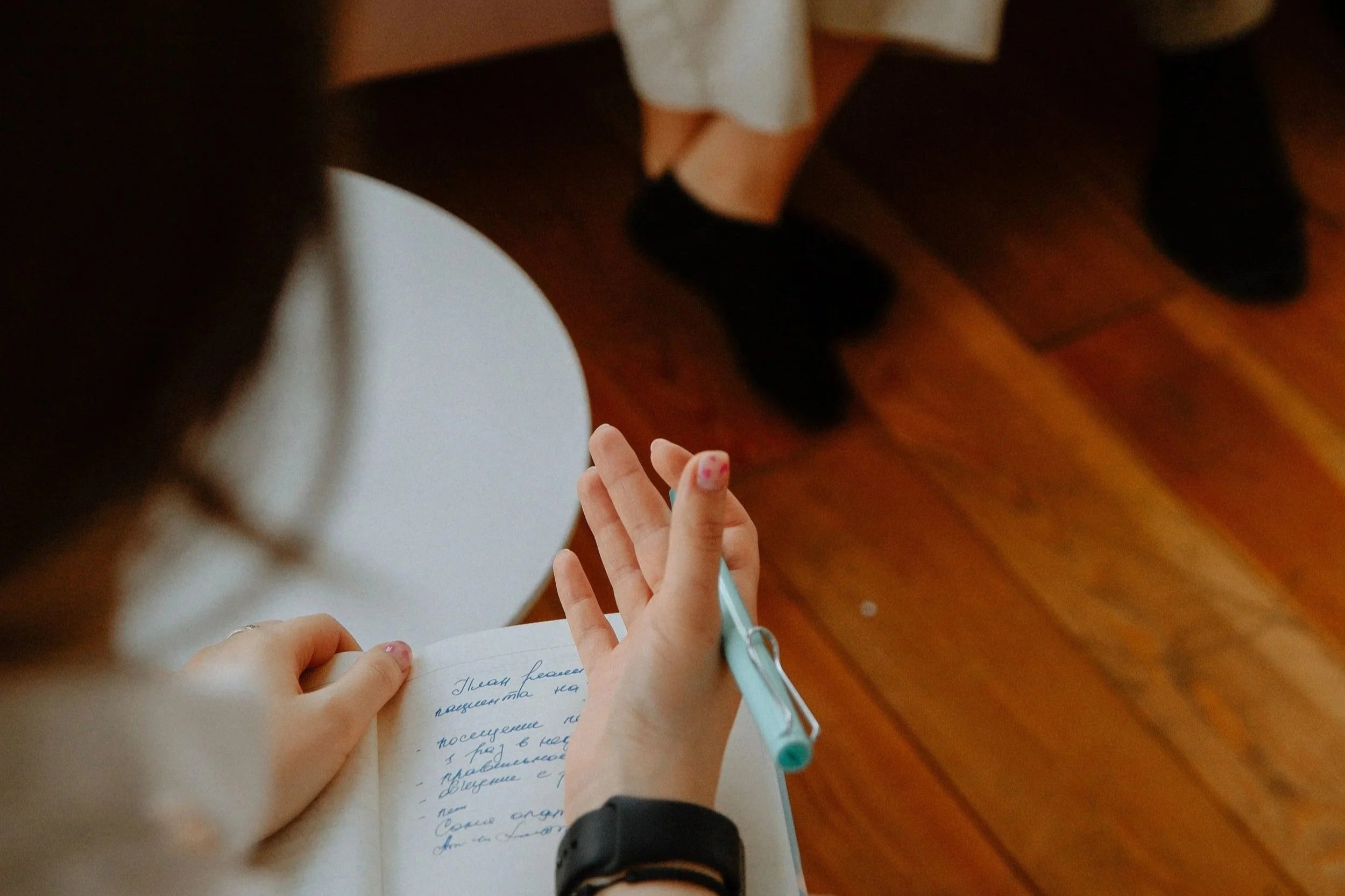 Une personne écrit dans un carnet avec un stylo turquoise, un enfant fait signe avec la main, en arrière-plan un adulte avec des chaussures noires sur un sol en bois.