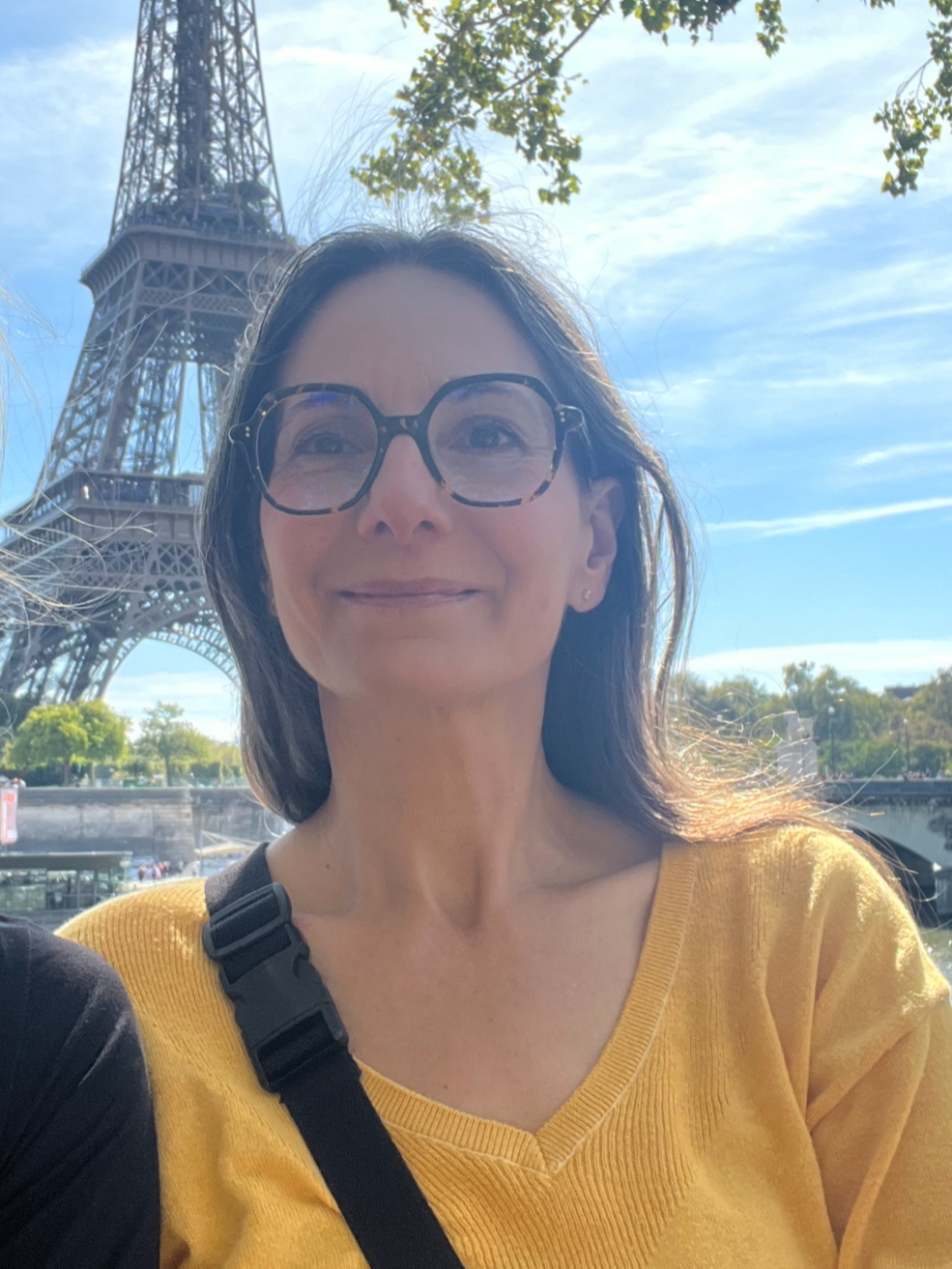 Femme portant des lunettes et un pull jaune, devant la Tour Eiffel à Paris, par une journée ensoleillée.