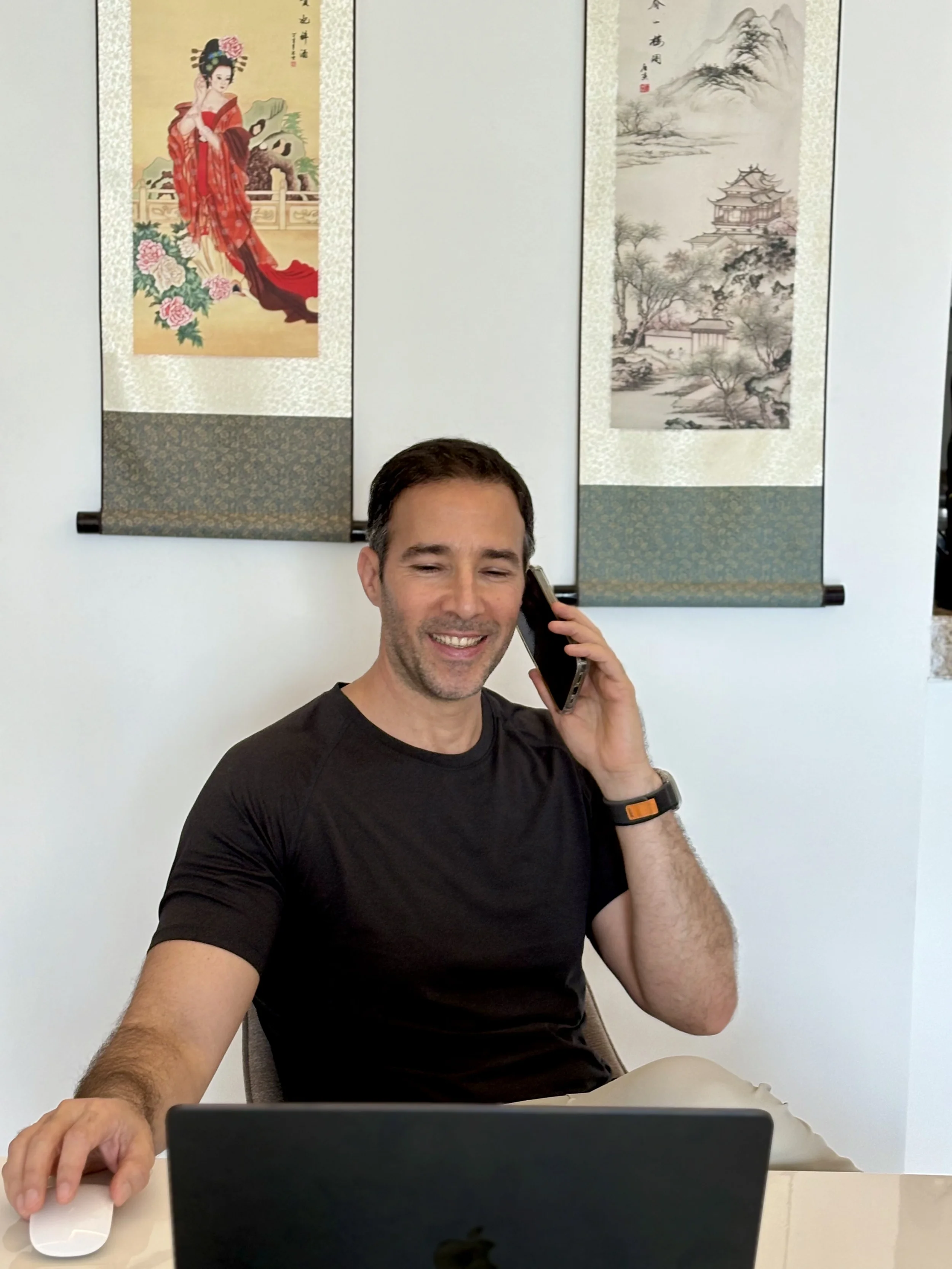 A man, health coach, Joe Sfeir,  smiling and talking on a cellphone while sitting at a desk with a computer mouse and a laptop, with traditional Asian scroll paintings on the wall behind him.