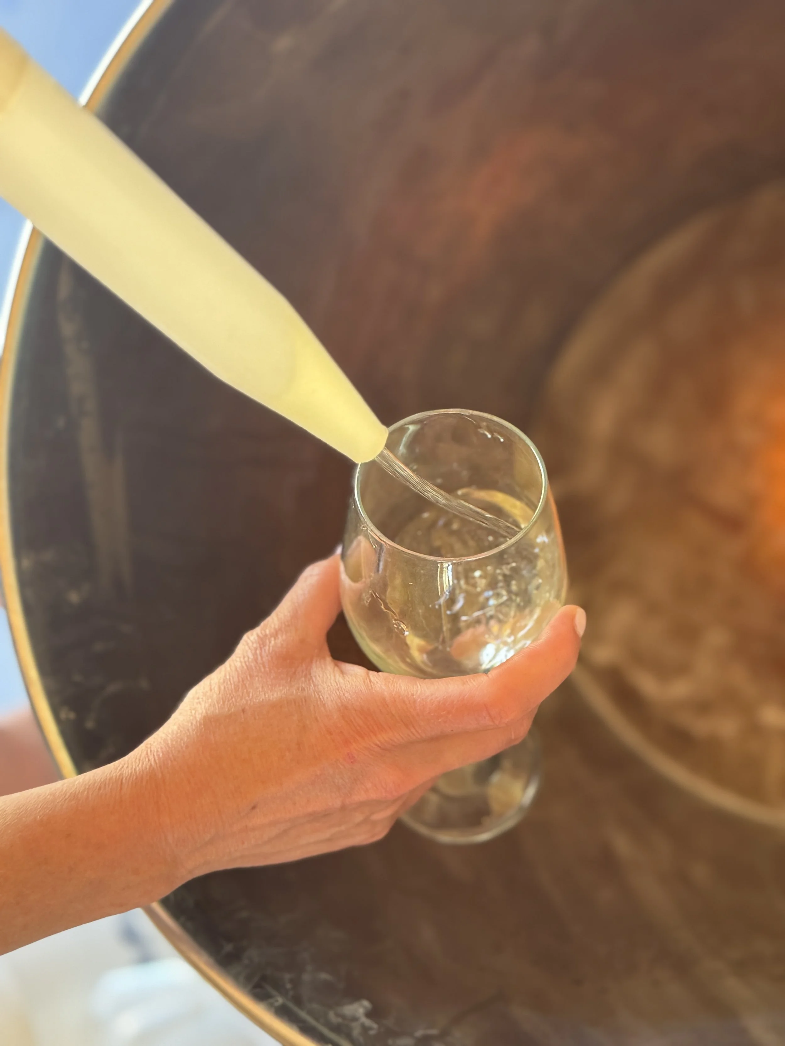 A hand holding a clear glass filled with a transparent liquid, with a yellow candle or tube being poured into it over a brown surface.