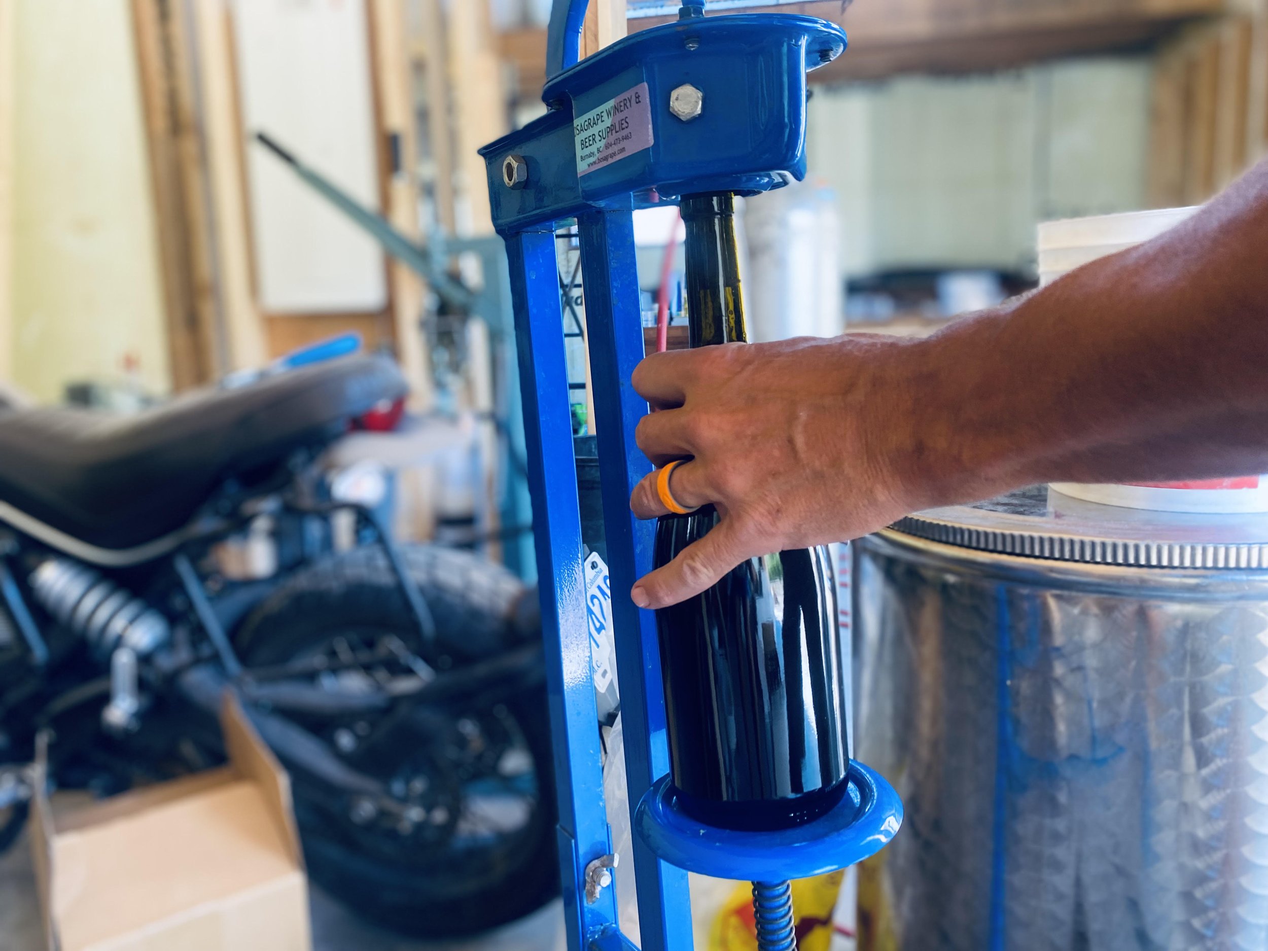 Close-up of a person's hand wearing two rings adjusting a blue metal lock or clamp in a workshop or construction area with wooden beams and tools in the background.