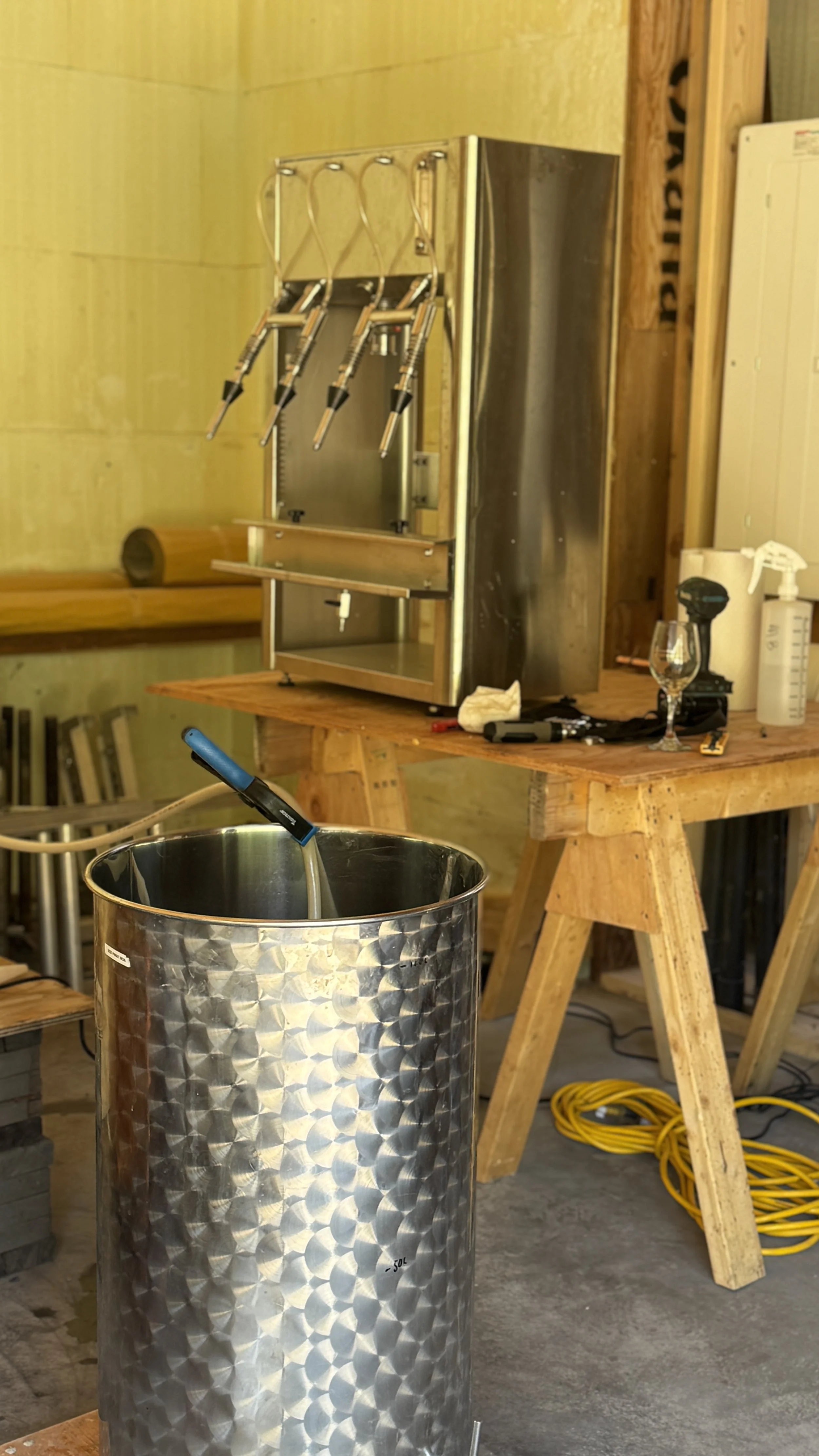 A metal beer can with a filled water pipe inside, a wooden worktable, a faucet with a glass, tools, and a large spray bottle on the table. In the background, there is a brewing machine with three nozzles.