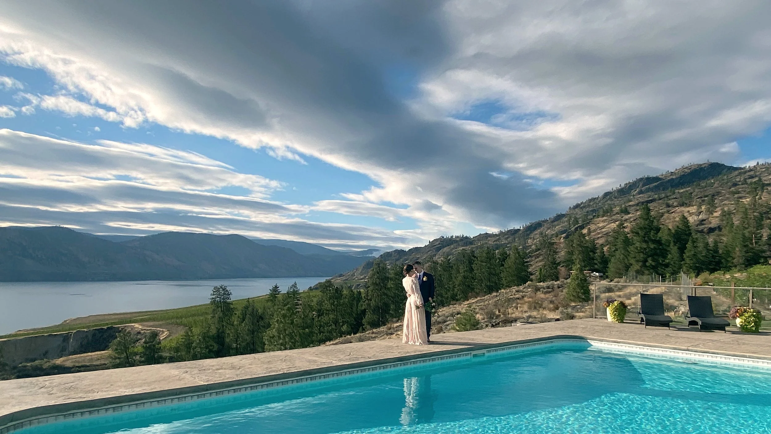 A couple in wedding attire stands by a scenic lake with mountains in the background, next to a swimming pool on a partly cloudy day.