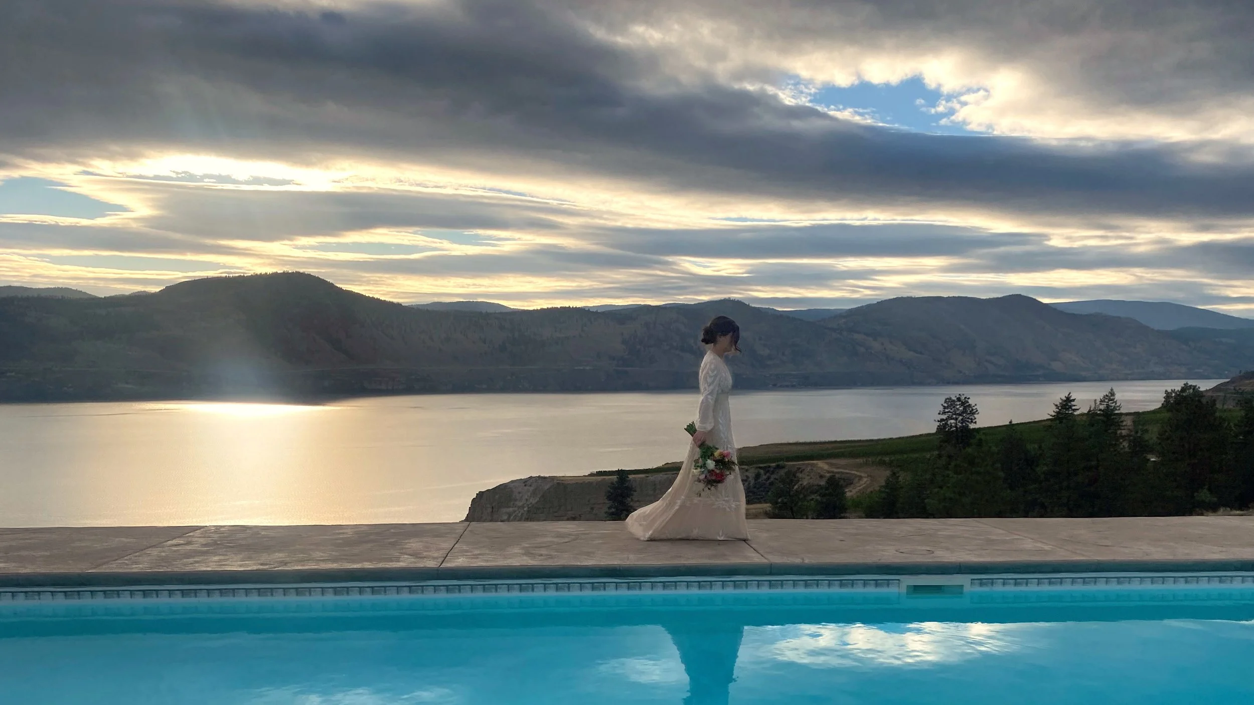 A woman in a white dress holding a bouquet of flowers stands outside near a body of water, with mountains and a cloudy sky in the background.