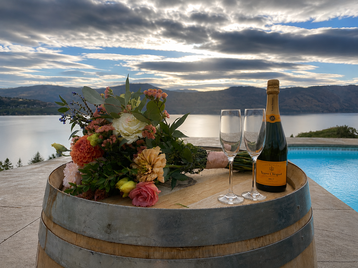 A bouquet of colorful flowers, a bottle of Veuve Clicquot champagne, and two champagne flutes on a wooden barrel, with a scenic lake and mountain landscape in the background during sunset.