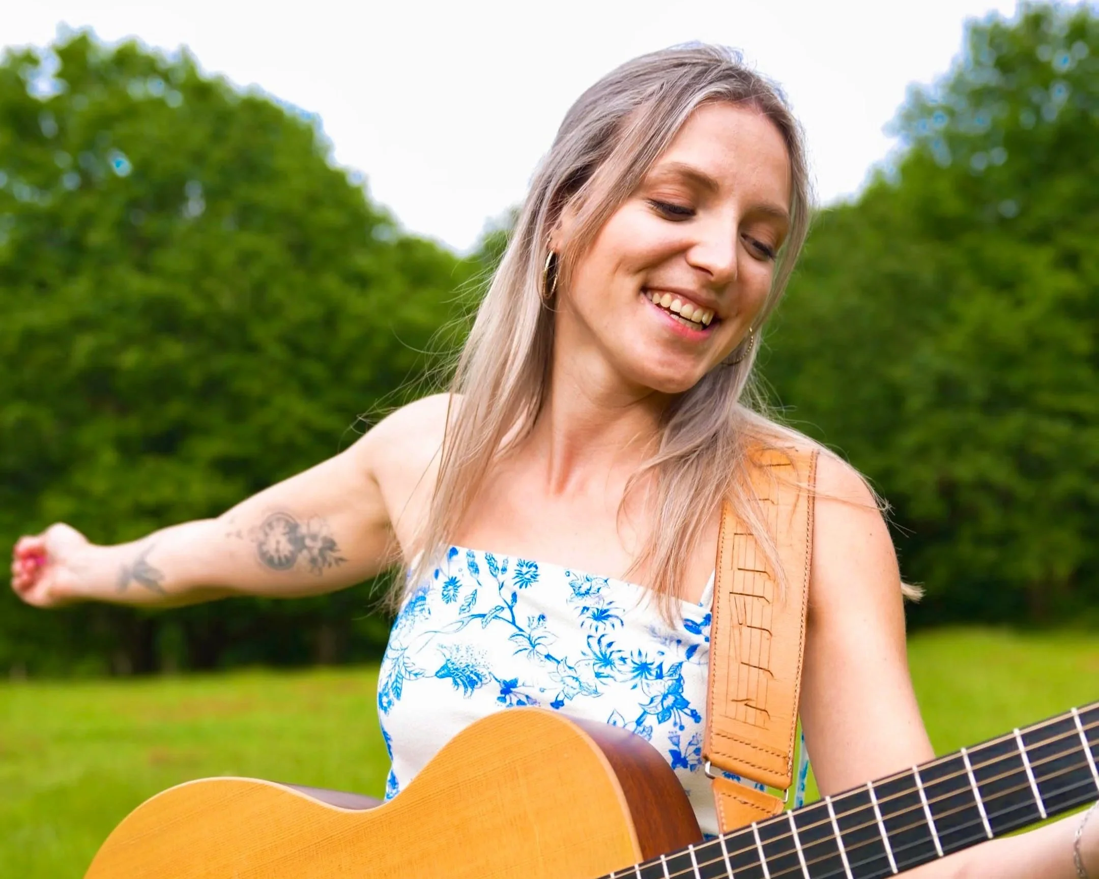 Young woman with blonde hair smiling and playing an acoustic guitar outdoors in a park with green trees in the background.
