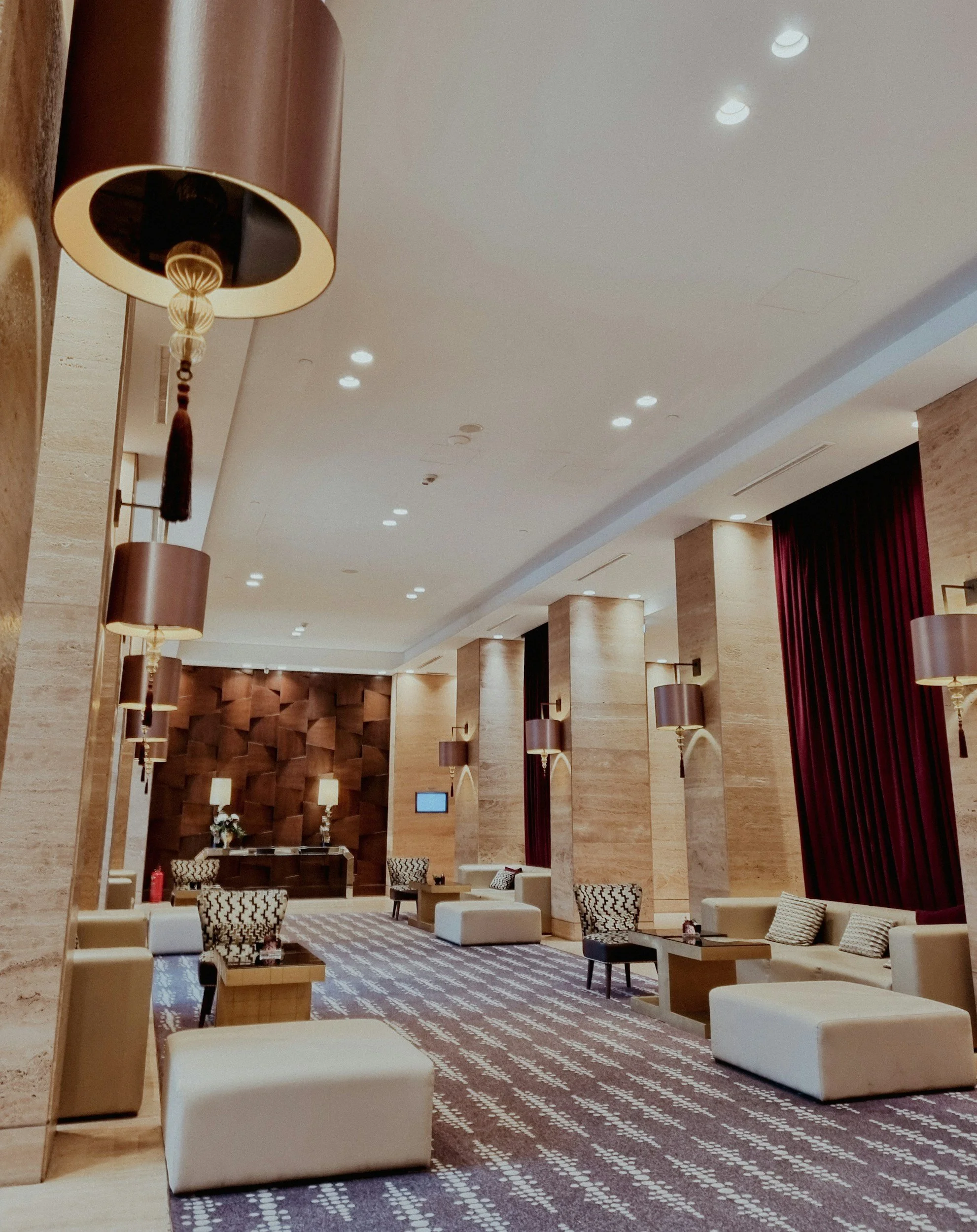Luxury hotel lobby featuring beige upholstered seating, patterned rugs, dark red curtains, beige walls, and contemporary light fixtures.