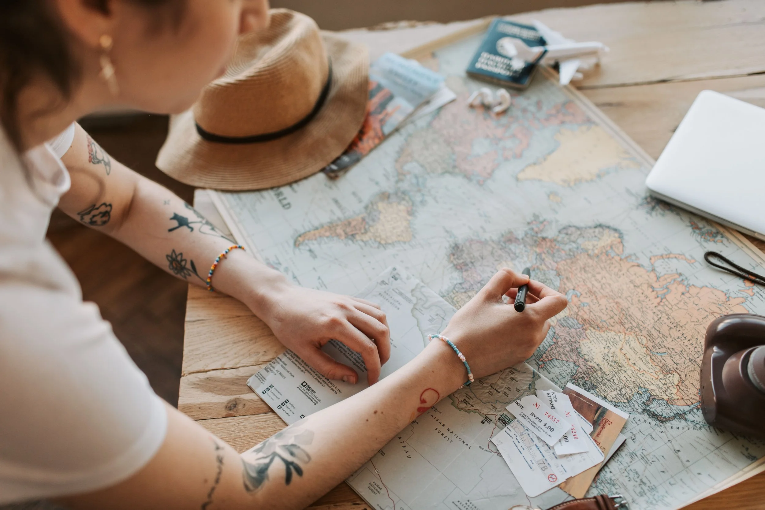 A person with tattoos on their arm sitting at a wooden table, preparing to travel. They have a straw hat, multiple travel tickets, a map, sunglasses, and a laptop on the table. The person is writing or drawing on the map.