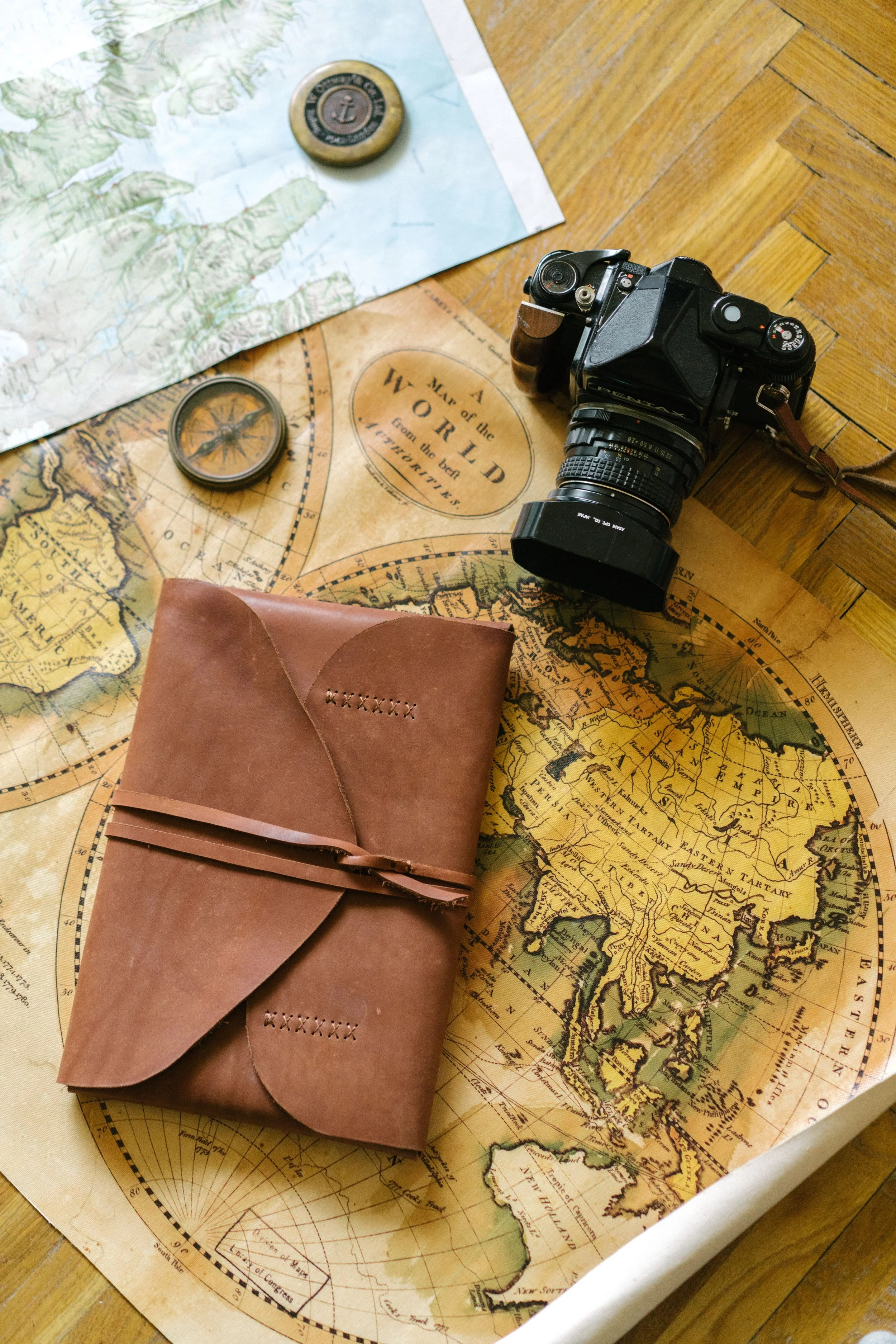 A vintage map on a wooden surface with a camera, a compass, a navigational token, and a brown leather wallet placed on and around it.