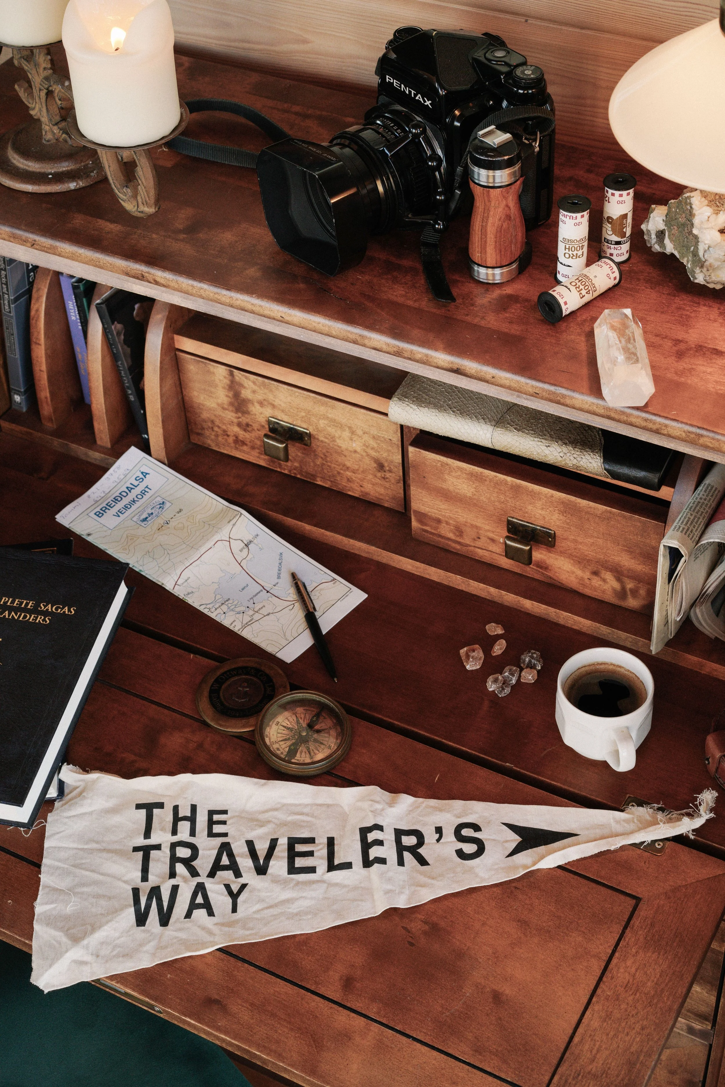 A wooden table with a candle, a camera, several film rolls, stones, a map, a mug of coffee, and a cloth with the words 'THE TRAVELER'S WAY' printed on it.