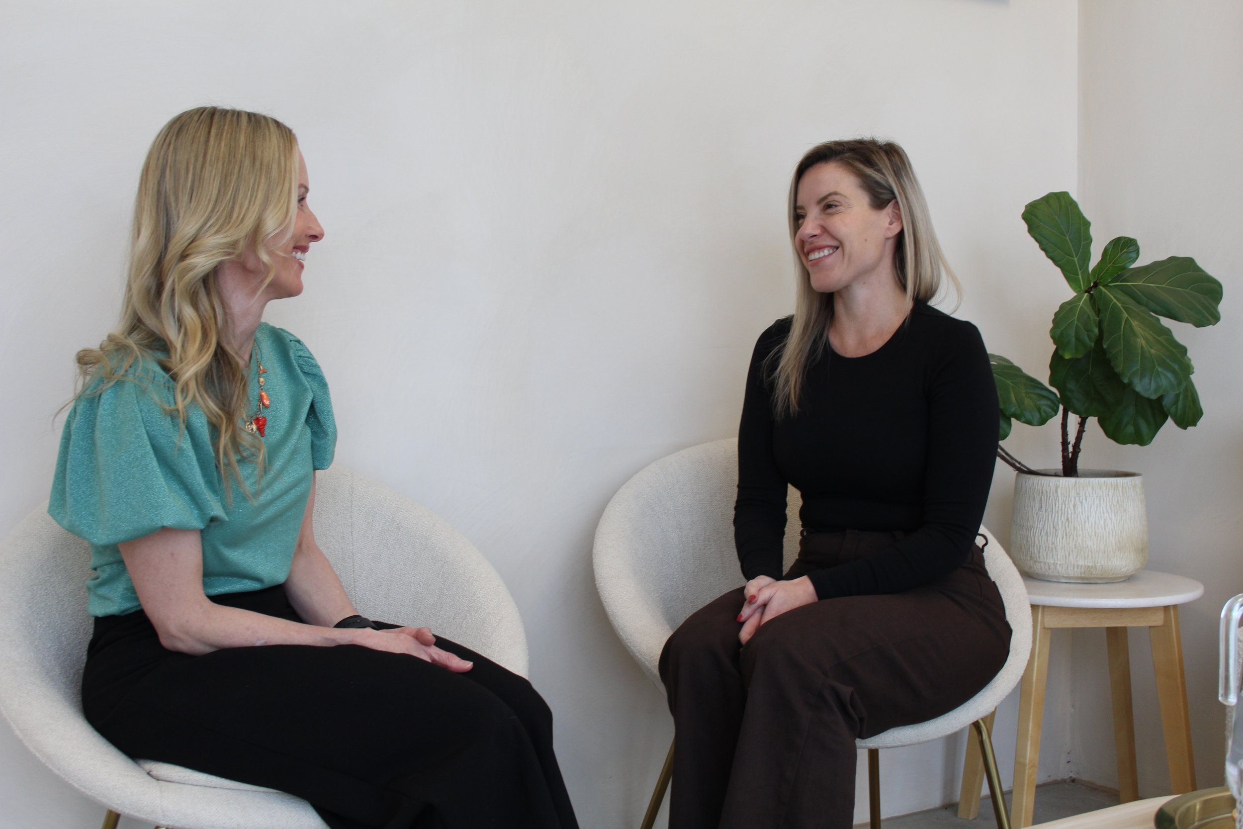 Two women sitting and smiling at each other in a bright, minimalist room with a large potted plant beside them.