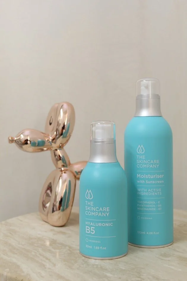 Two bottles of skincare products from The Skincare Company on a wooden surface, with a metallic balloon sculpture shaped like a dog in the background.
