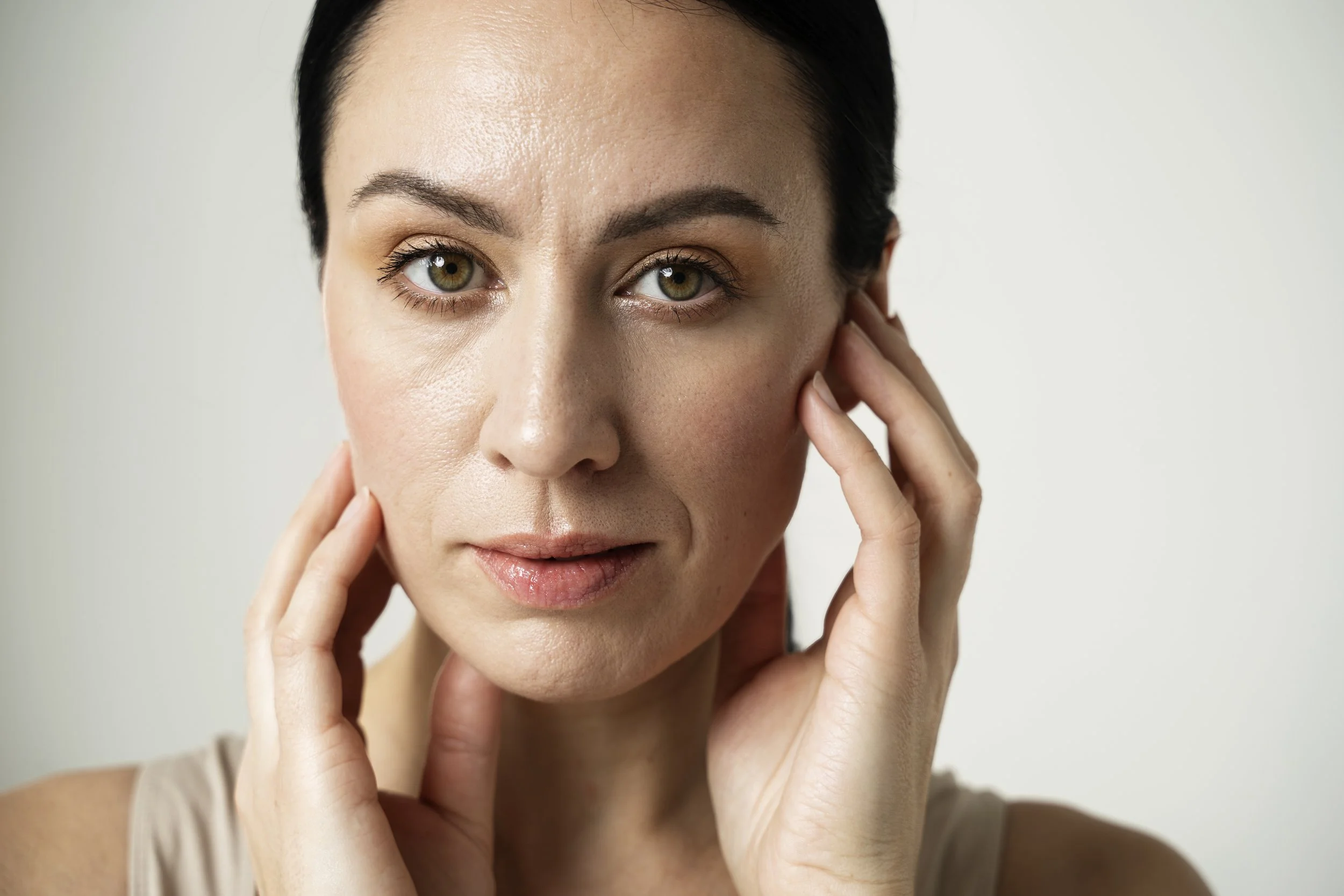 A close-up of a woman with dark hair touching her face, showing clear skin and light makeup, against a plain white background.