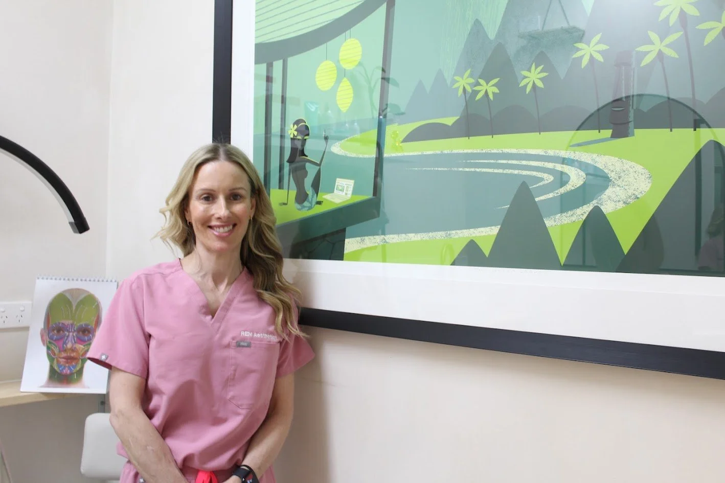 A woman in pink scrubs standing in a clinical room with a large framed illustration of a stylized nature scene on the wall behind her, and a colorful face chart on a desk to her side.