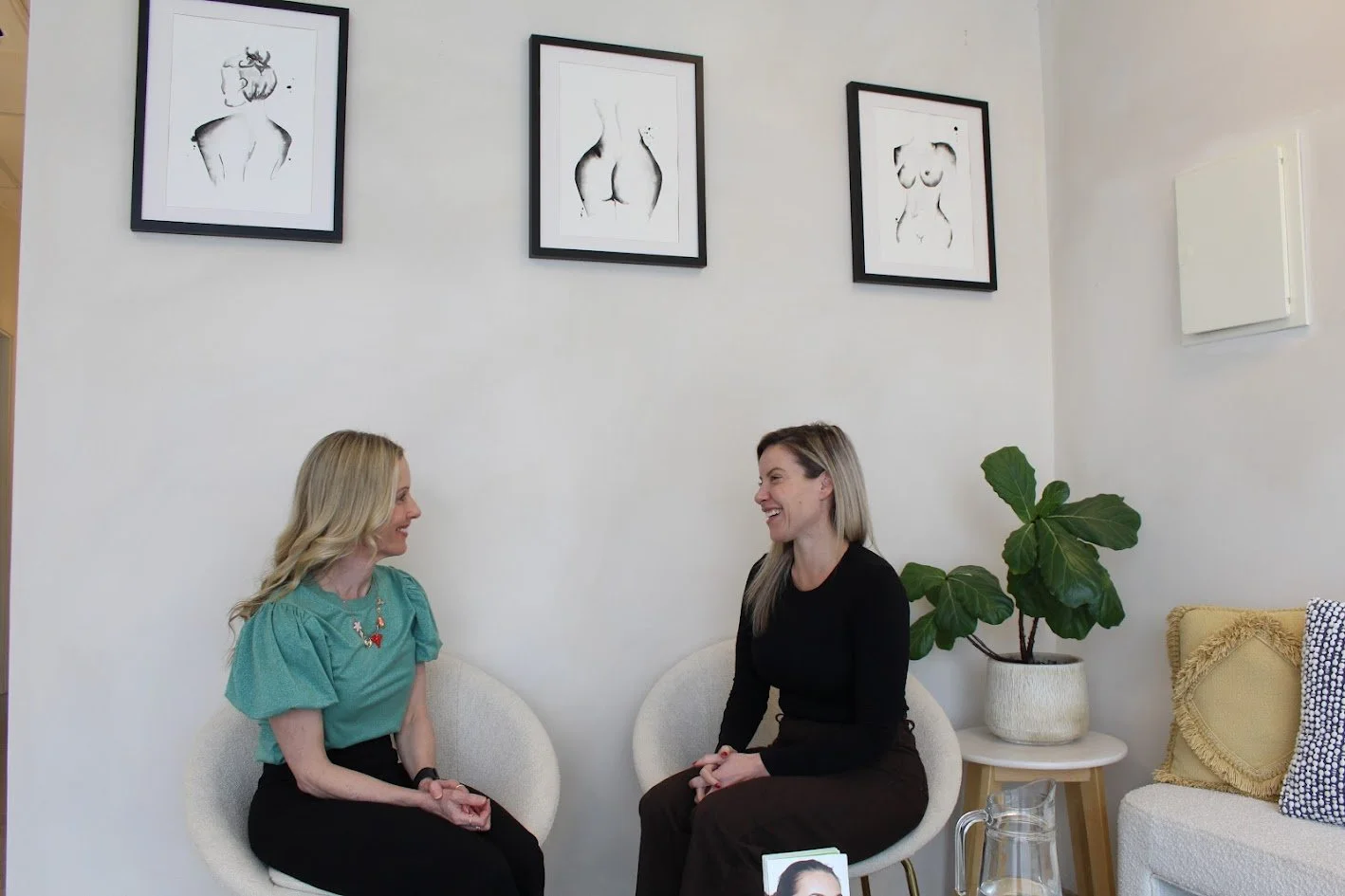 Two women sitting on white chairs, talking and smiling in a bright room with framed art of female figures on the wall and a large potted plant.