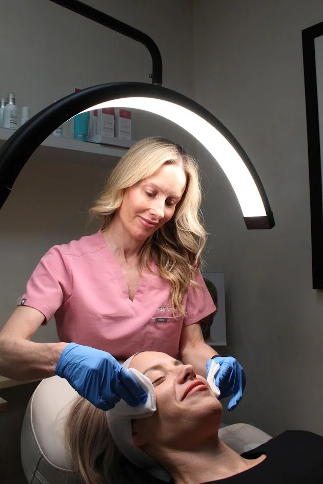 A beauty specialist wearing a pink uniform and blue gloves is performing a facial treatment on a woman lying in a skin care clinic, illuminated by a bright, curved light.