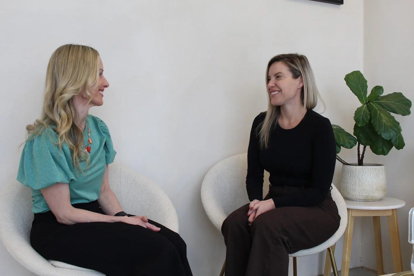 Two women sitting and smiling at each other in a room with white walls, a potted plant on a side table, and modern chairs.