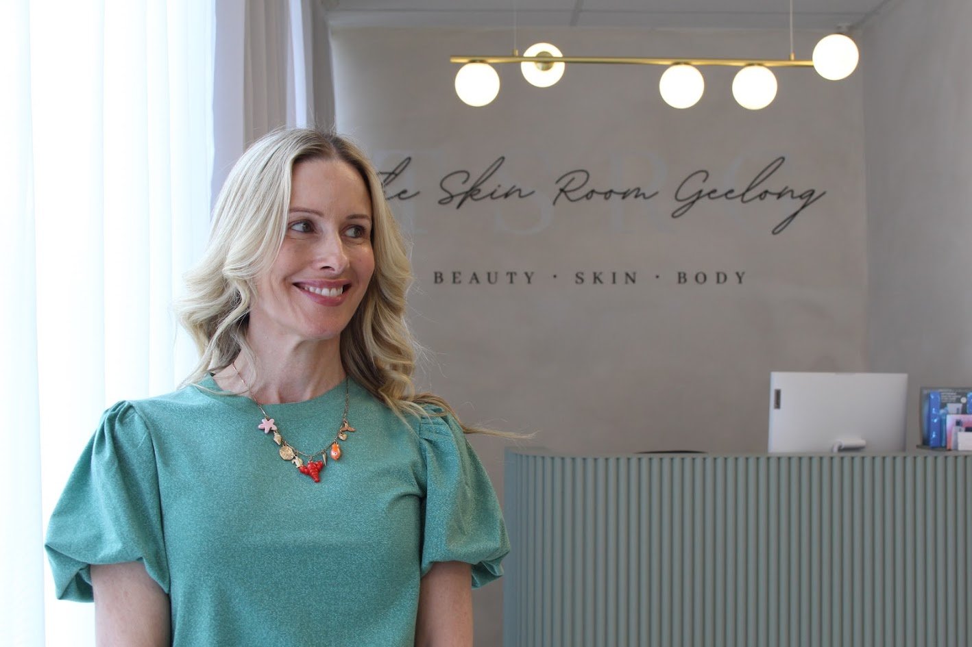 A woman with blonde hair smiling inside a skin care clinic, behind a reception desk, with a sign on the wall that reads 'The Skin Room Geelong' and 'Beauty Skin Body' under it.