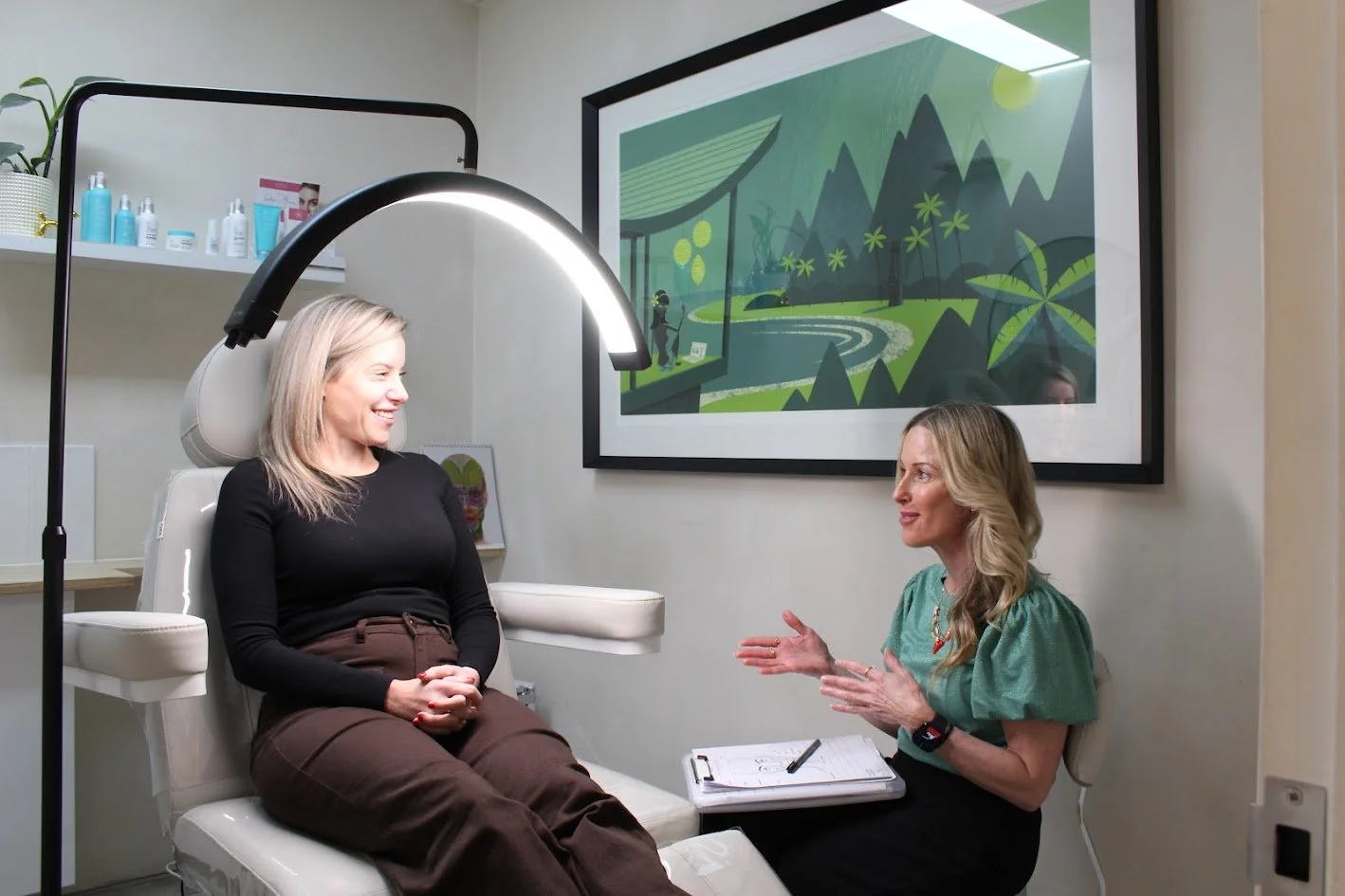 A woman sitting in a medical examination chair in a doctor's office, talking to a healthcare professional. The professional is gesturing with her hands and has a clipboard on her lap. There are shelves with skincare products and a framed art piece wi