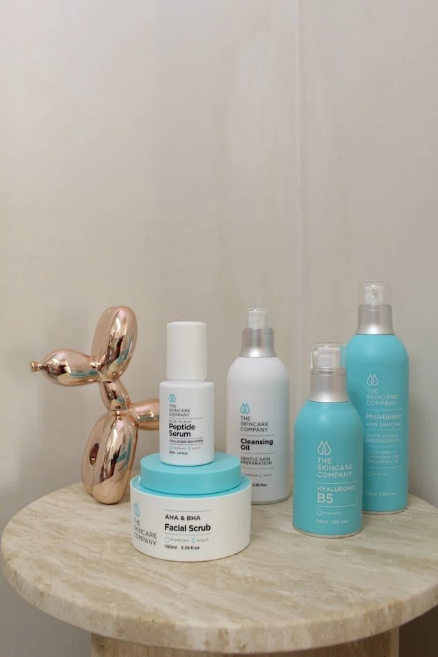 Skincare products on a round wooden table, including facial scrub, peptide serum, cleansing oil, and moisturizers, with a gold decorative balloon in the background.