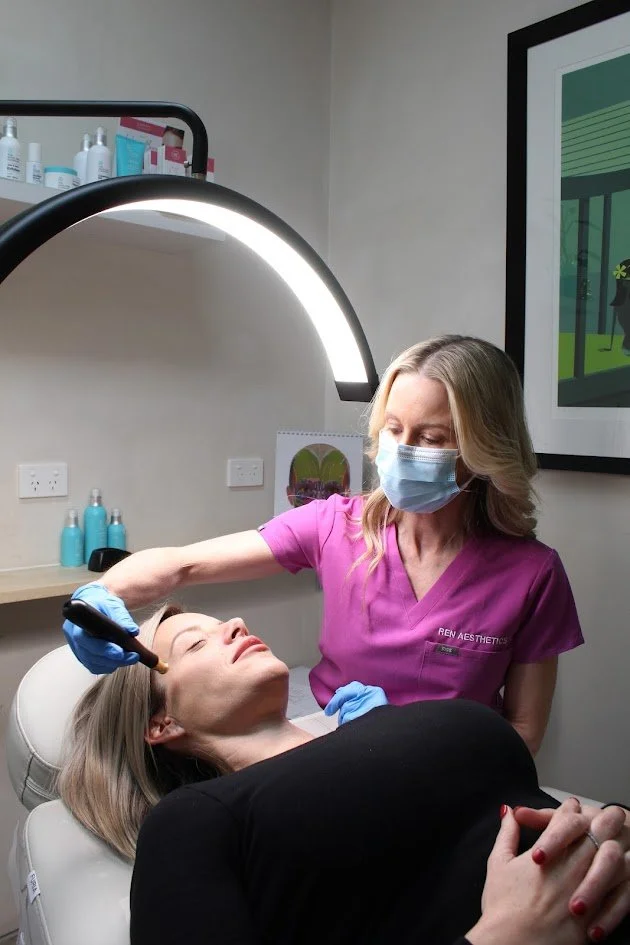 A woman receiving a cosmetic or dermatological treatment from a healthcare professional in a clinic. The professional is wearing a pink uniform and a face mask, while the patient is lying on a clinic bed under a bright light.