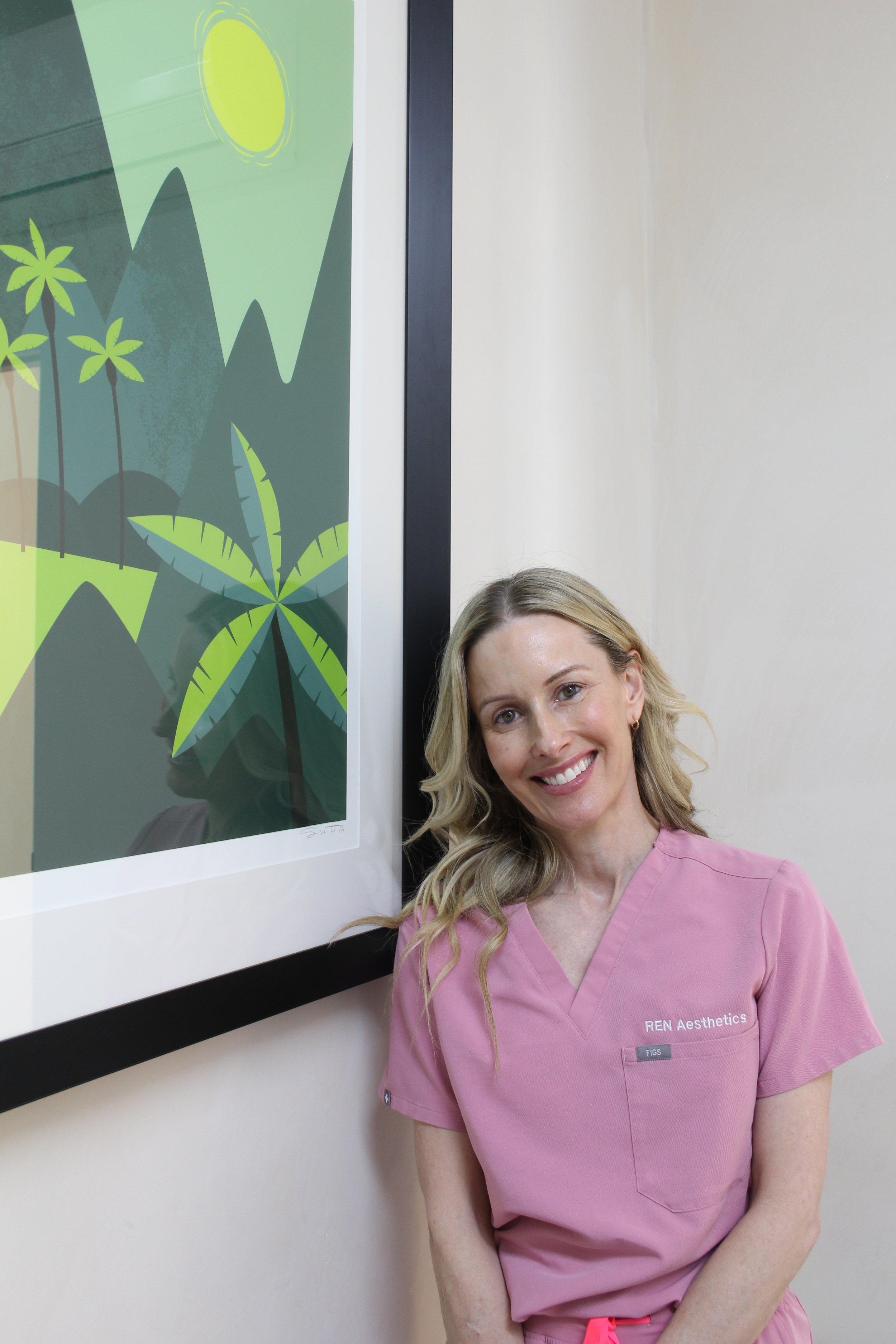 A smiling woman in pink medical scrubs standing beside framed artwork of stylized green and black mountains, palm trees, and a yellow sun.
